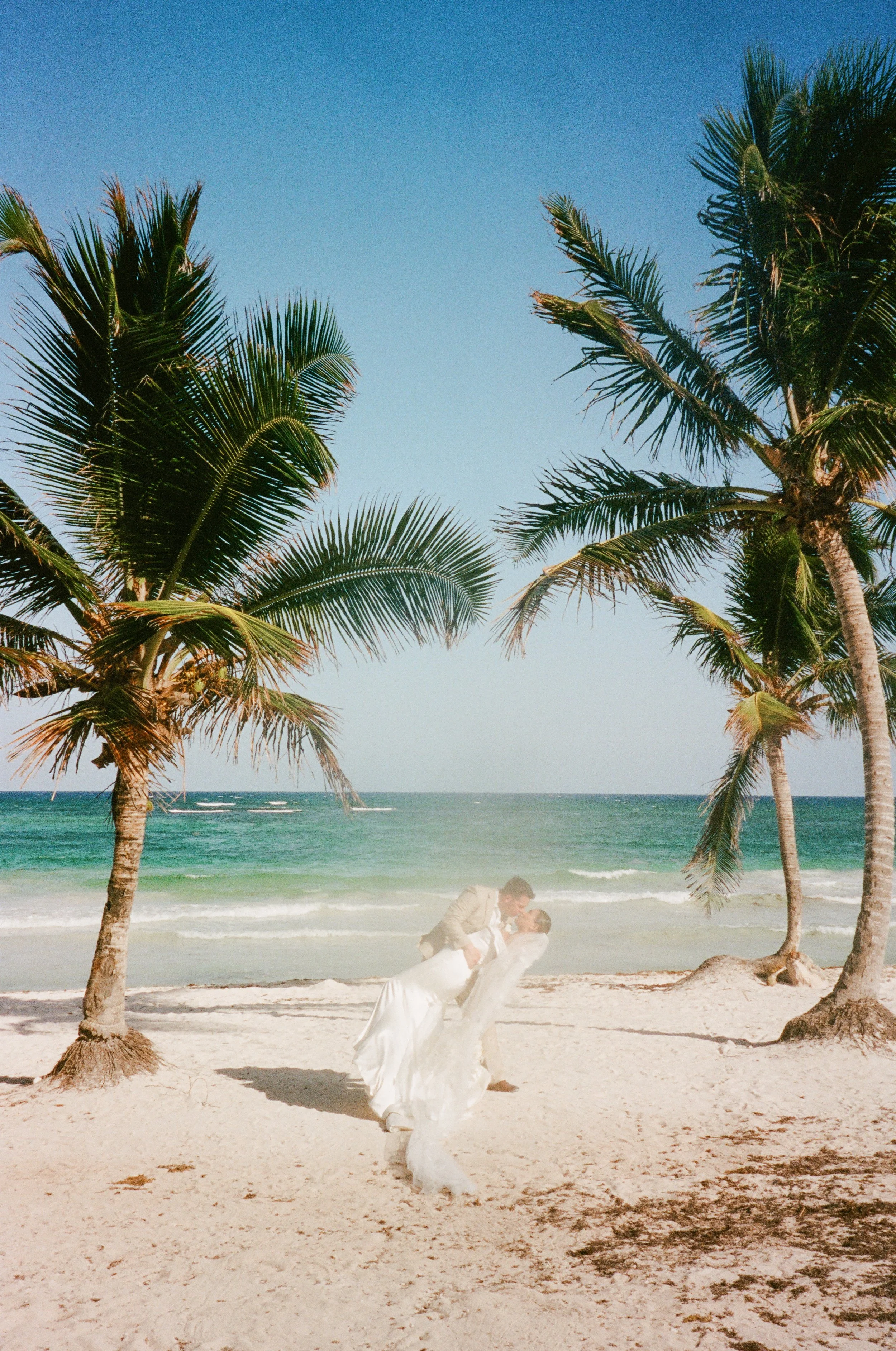 A man and woman on a beach under palm trees, with the man dipping the woman in a dance pose, and the ocean and sky in the background.