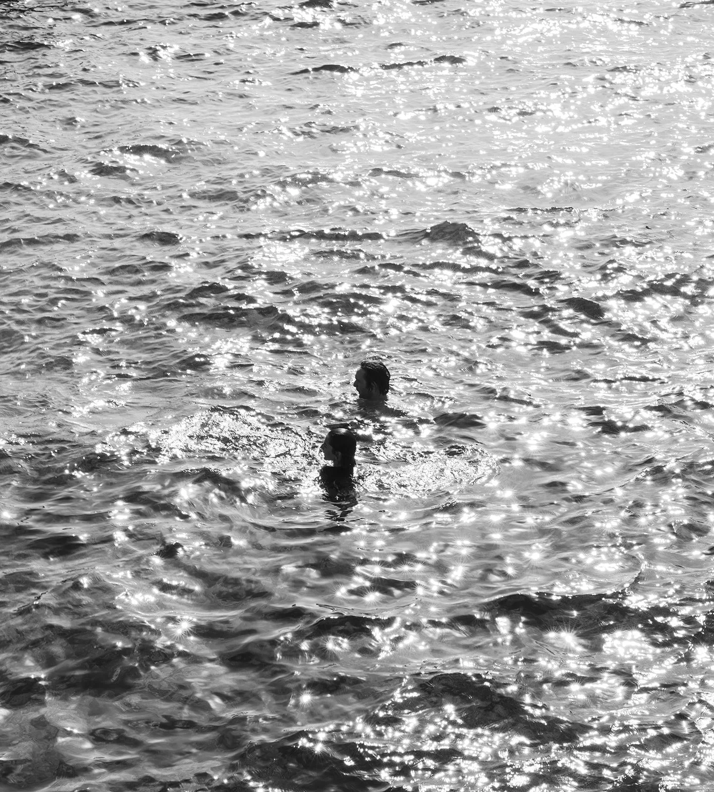 Two people swimming in a sunlit body of water, with reflections creating bright, sparkling effects on the surface.