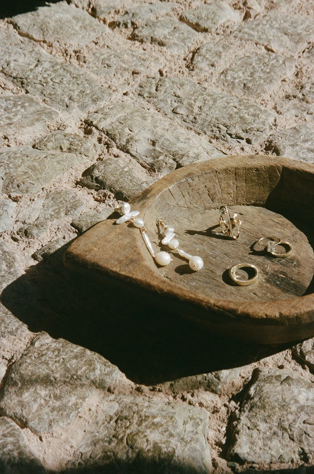 A wooden bowl on a rocky surface holding jewelry including pearl earrings and a pearl necklace.