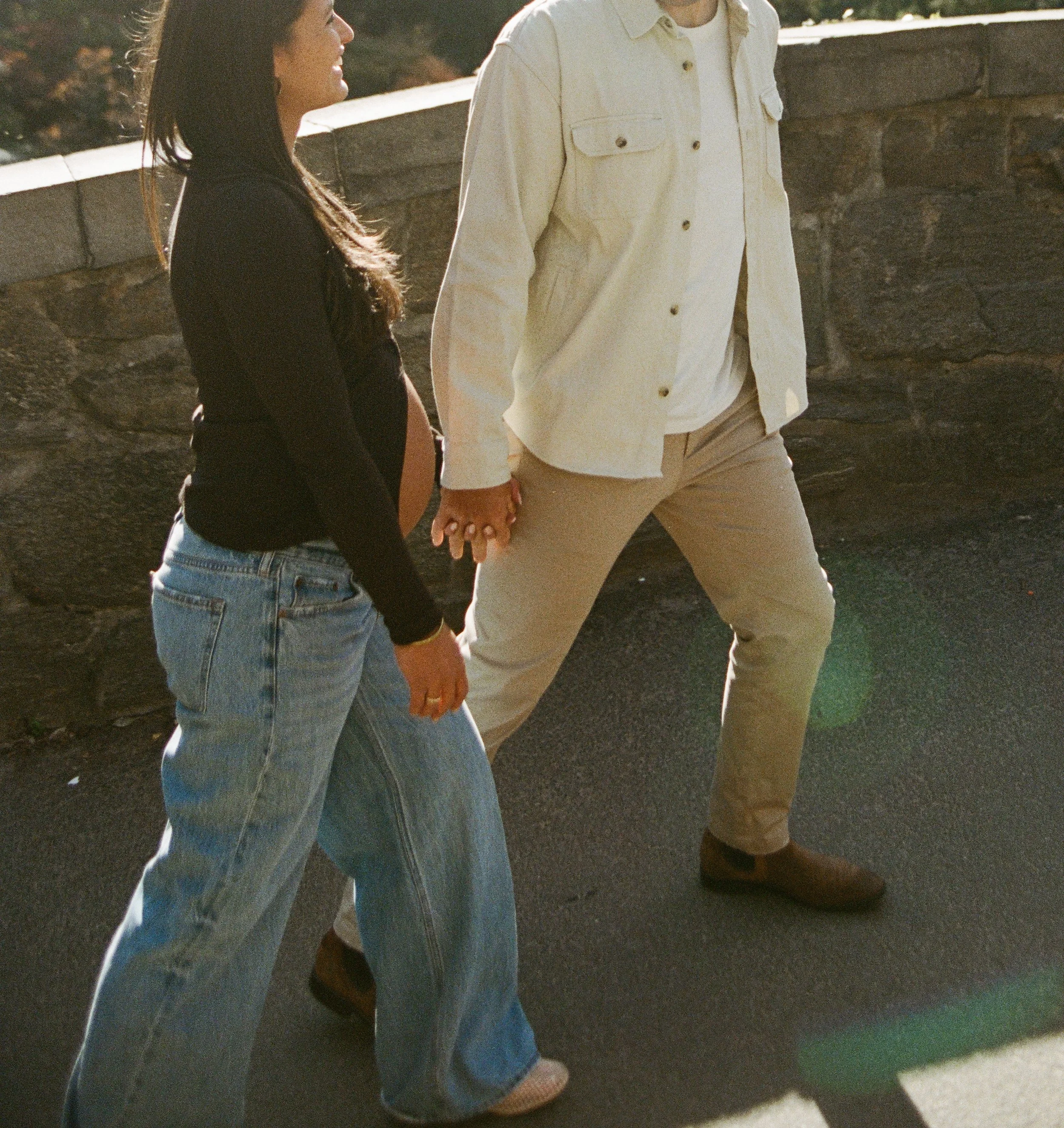 A couple holding hands and walking outdoors near a stone wall on a sunny day.