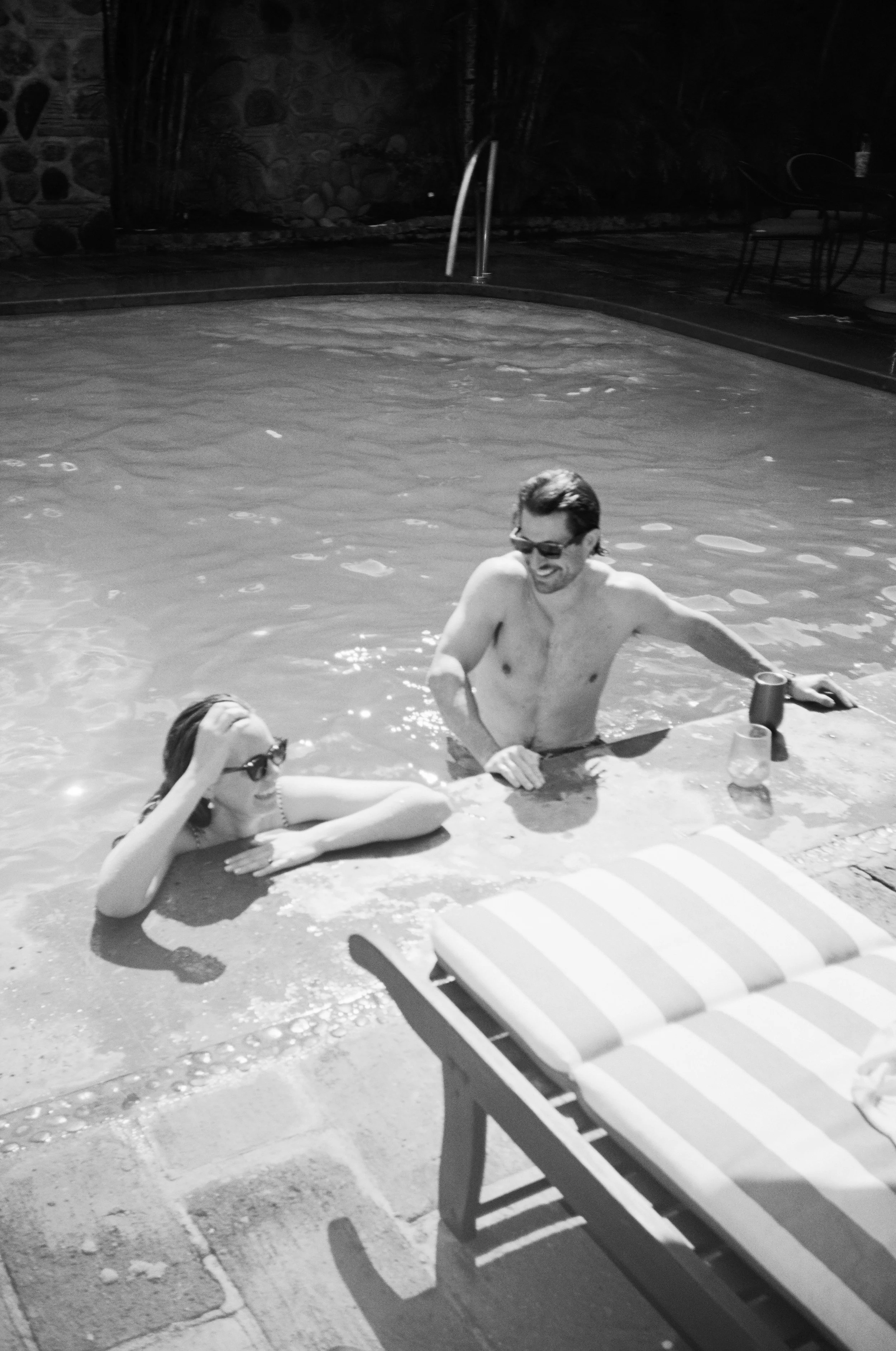 A man and woman relaxing in a swimming pool during daytime, with a lounge chair in the foreground and outdoor furniture on the patio.