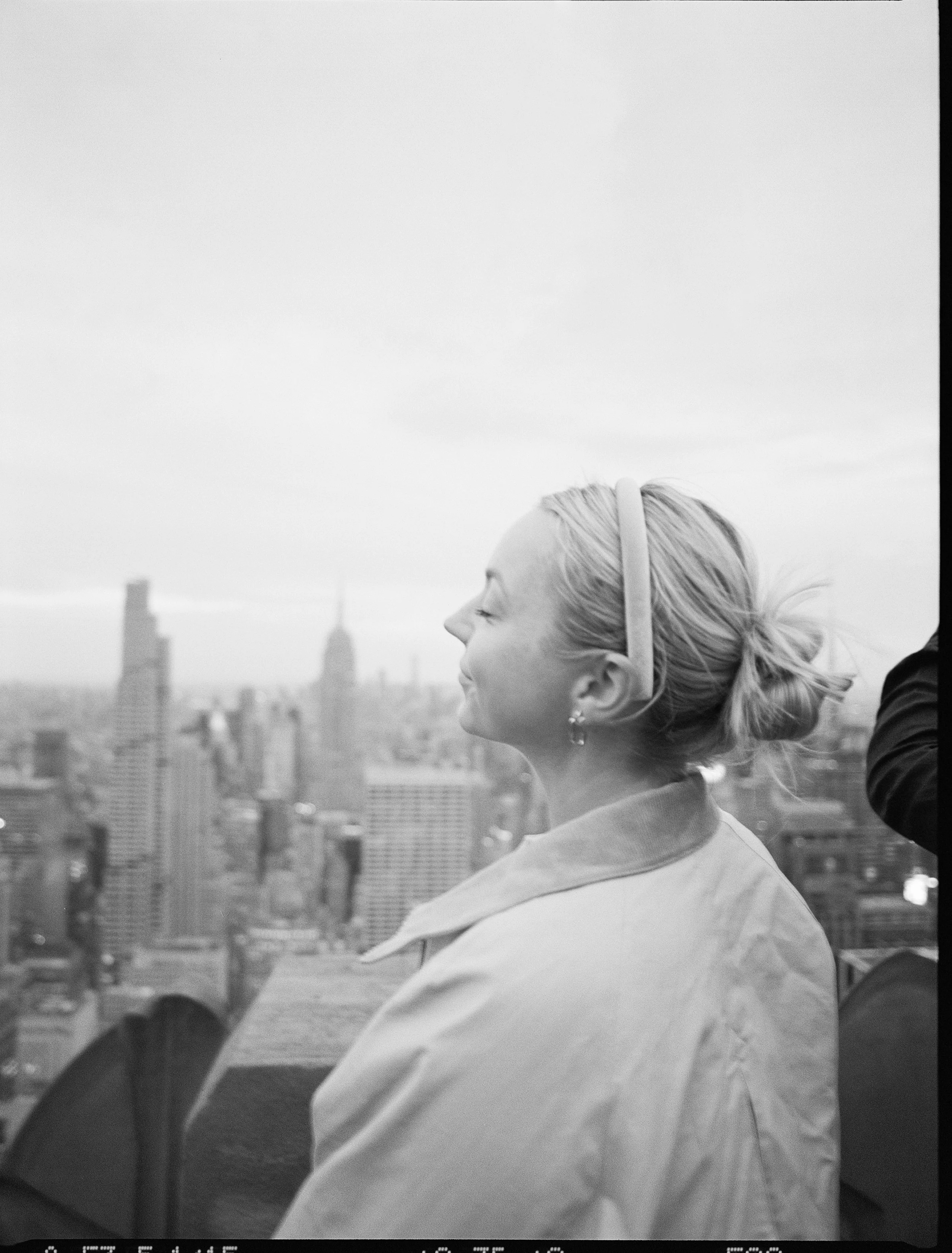 Grayscale photo of a woman with blond hair tied back, smiling with her eyes closed, wearing a light-colored jacket, standing outdoors on a rooftop with city buildings in the background.
