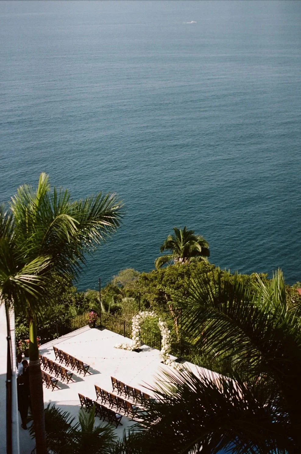An outdoor wedding setup on a terrace overlooking the ocean, surrounded by palm trees and greenery.