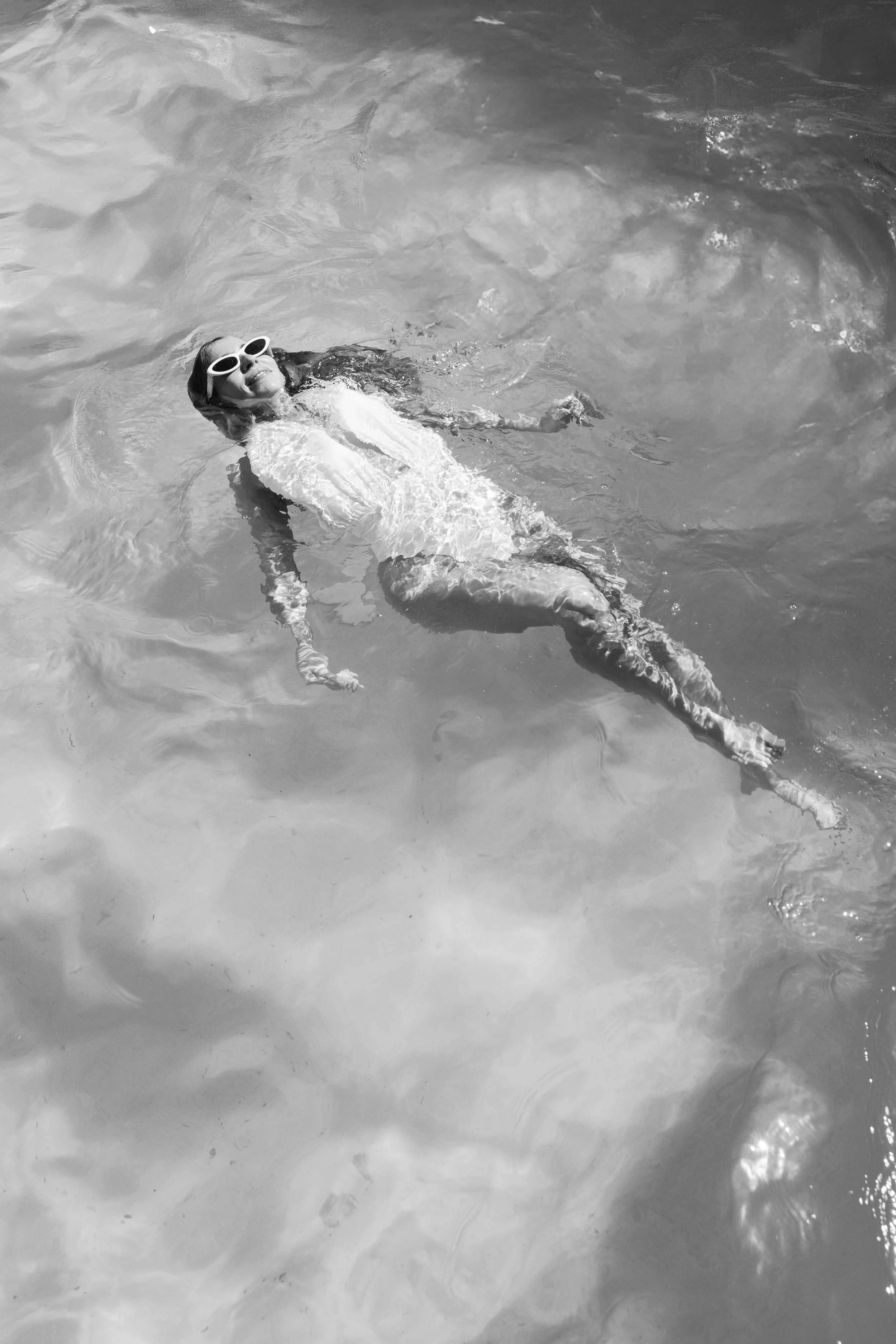 A woman floating in water, wearing white sunglasses and a white shirt, with her eyes closed and smiling.