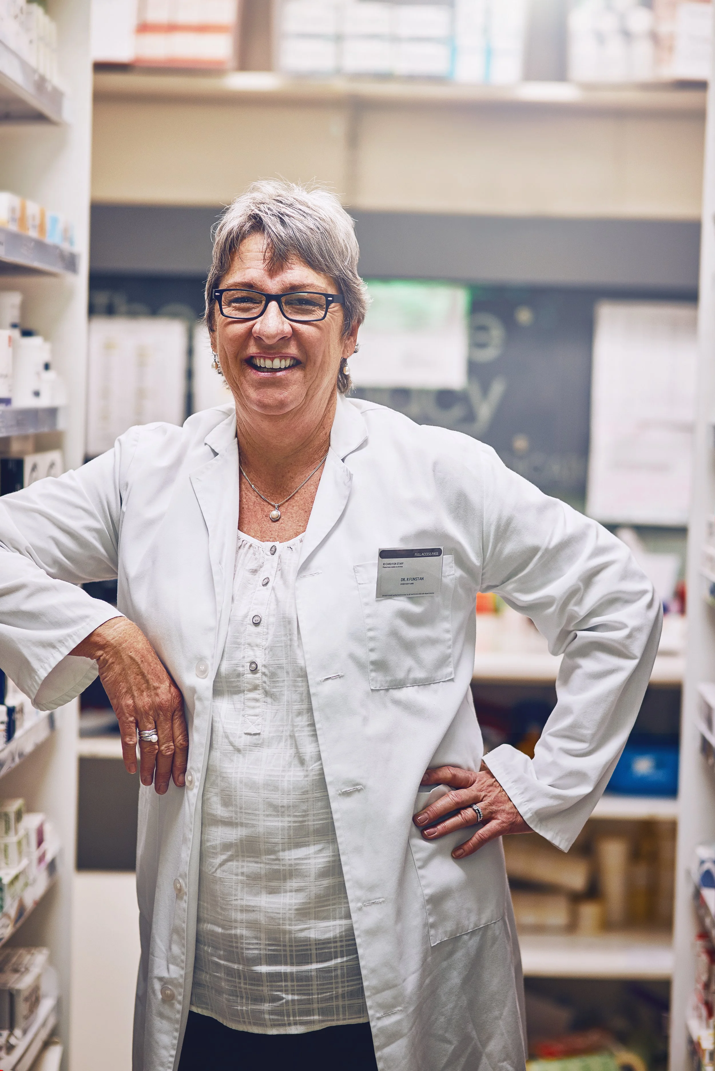 A smiling older woman with short gray hair and glasses, wearing a white lab coat and standing in a pharmacy aisle.