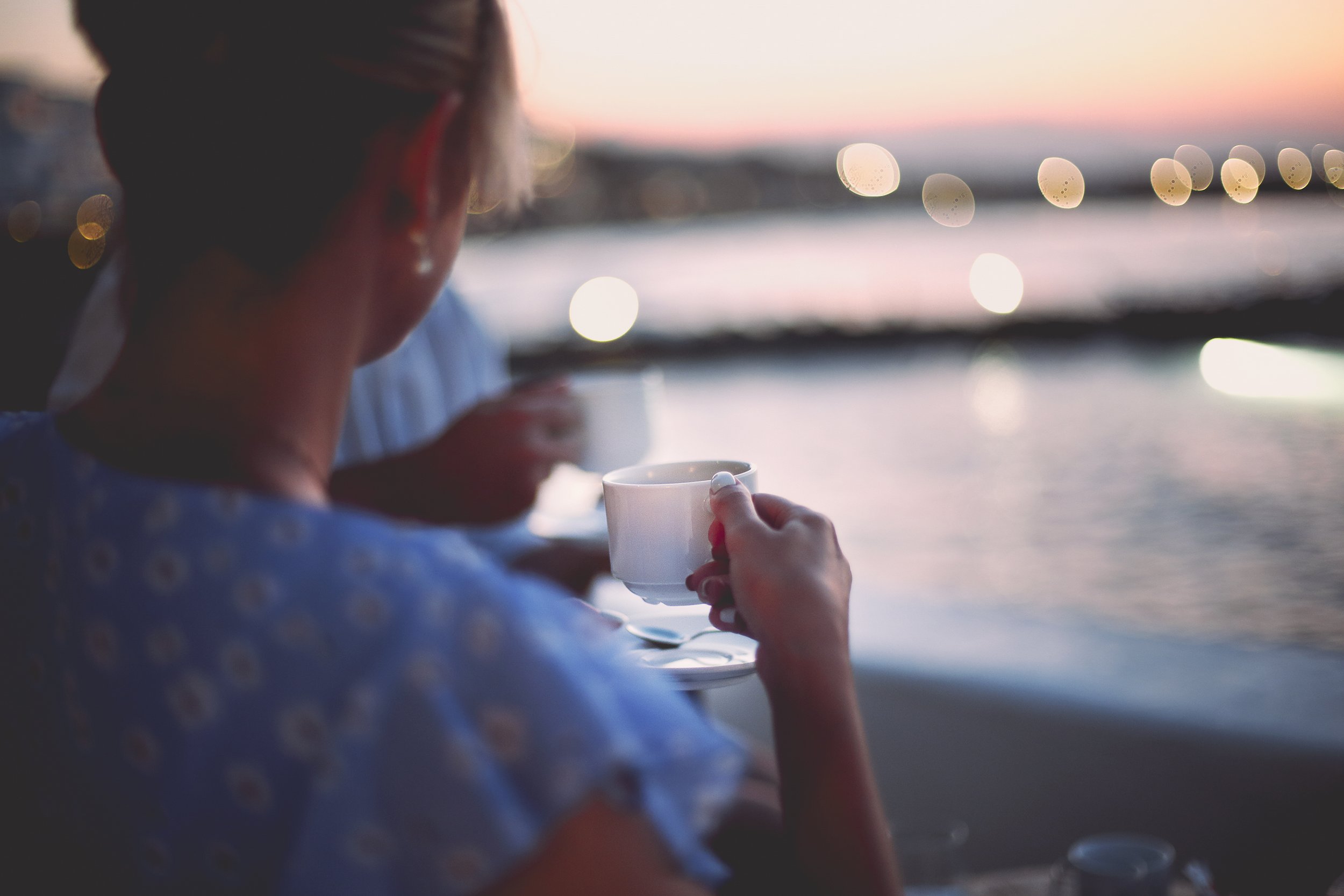 A woman with light brown hair, wearing a blue patterned shirt, holding a white coffee cup while sitting outdoors near a body of water during sunset, with blurred city lights in the background.