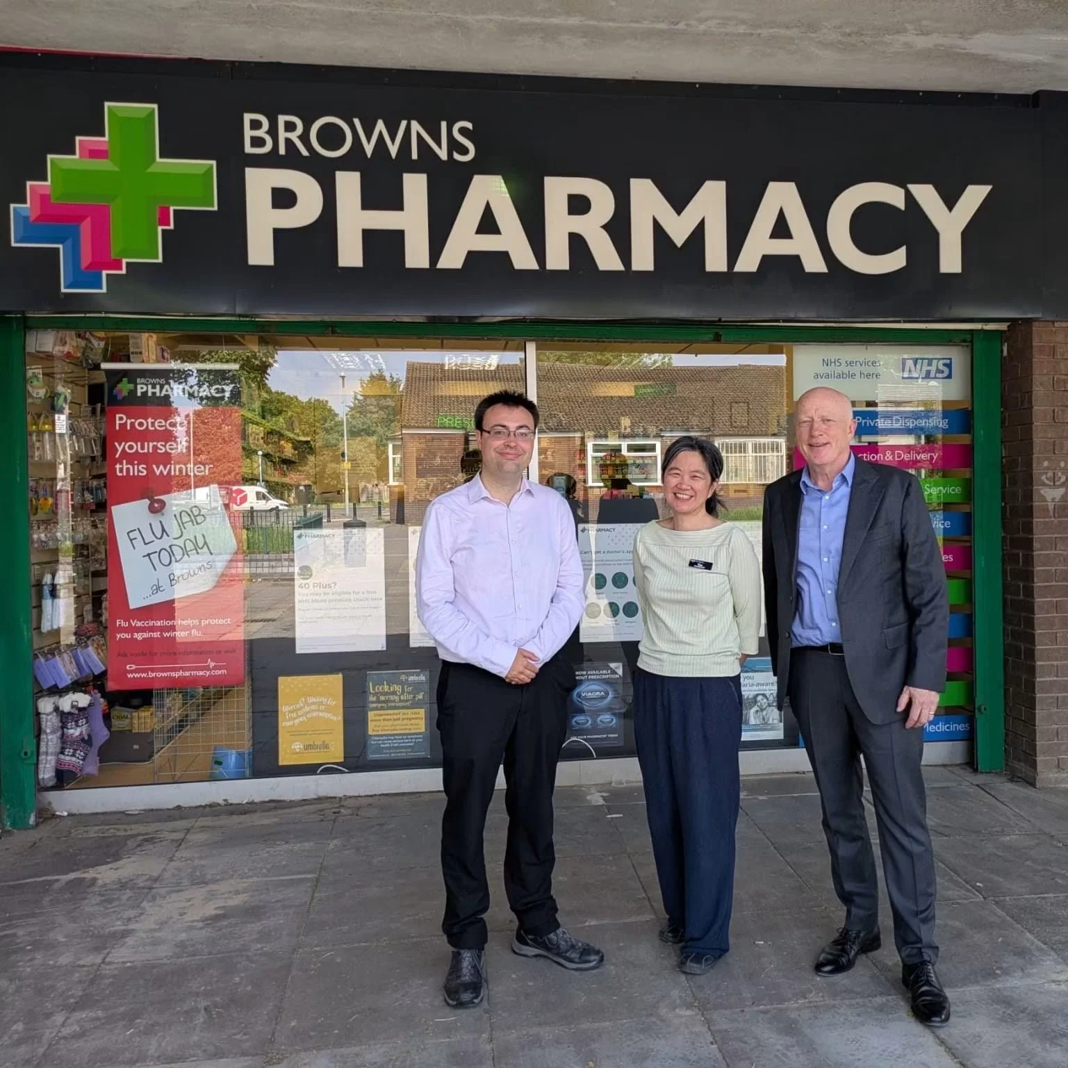 Three people standing outside a pharmacy with the sign "Browns Pharmacy." There are promotional posters in the window, including a red one about flu vaccines, and the background shows a residential neighborhood.