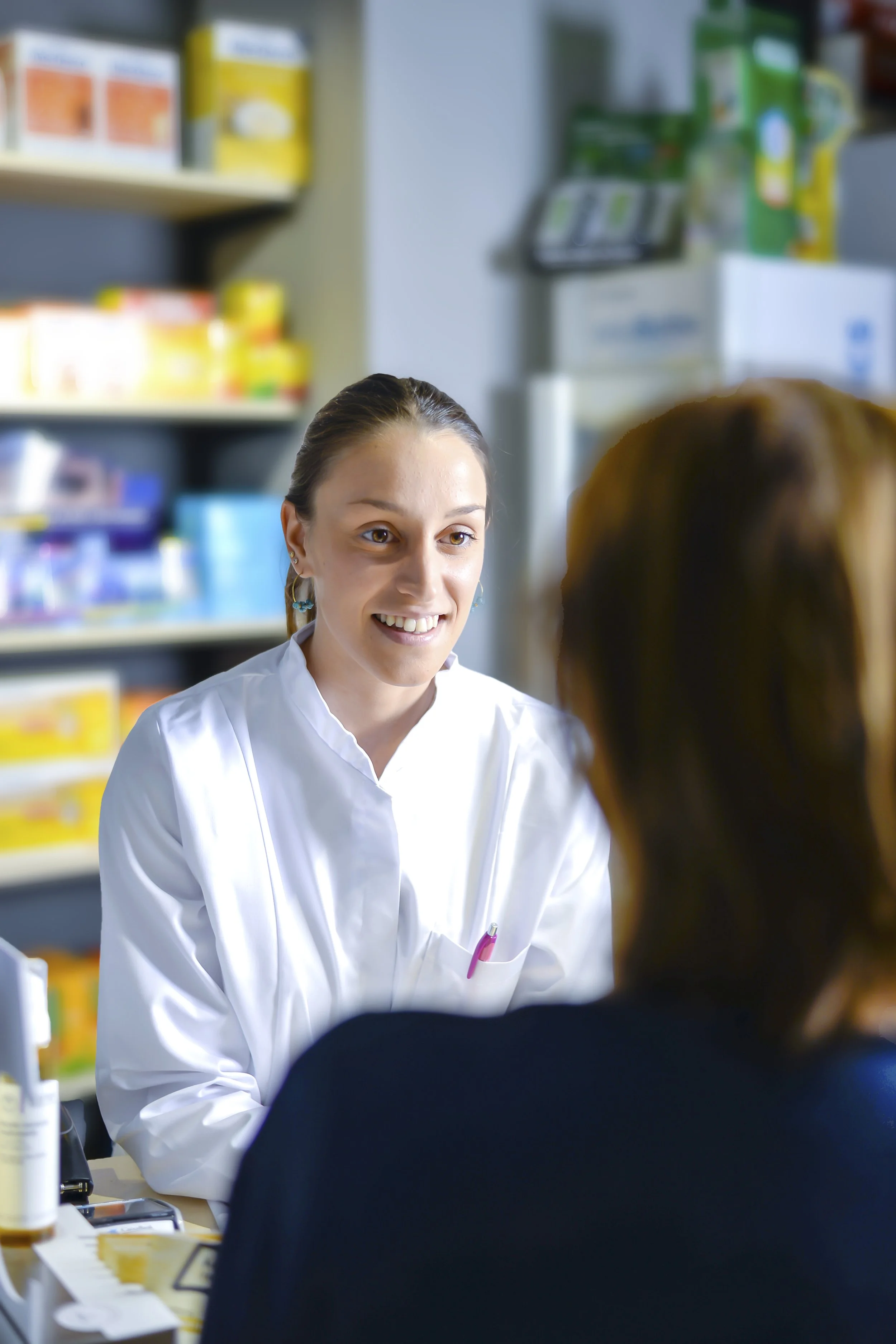 A young woman with brown hair and earrings, wearing a white lab coat, is smiling and talking to another person in a pharmacy or medical setting.
