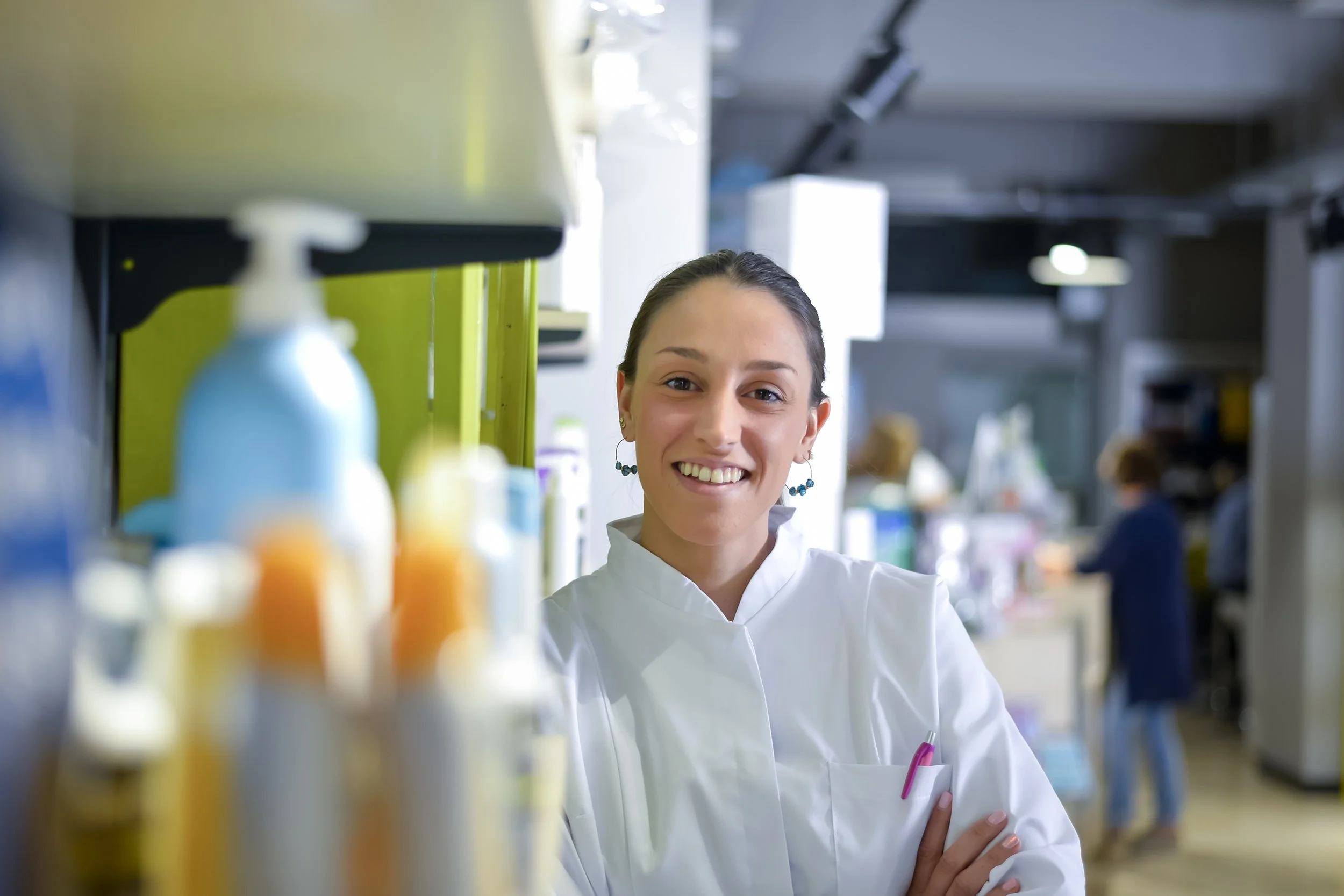 A female scientist or researcher in a laboratory setting, wearing a white lab coat, smiling, with various bottles and laboratory equipment in the foreground and background.