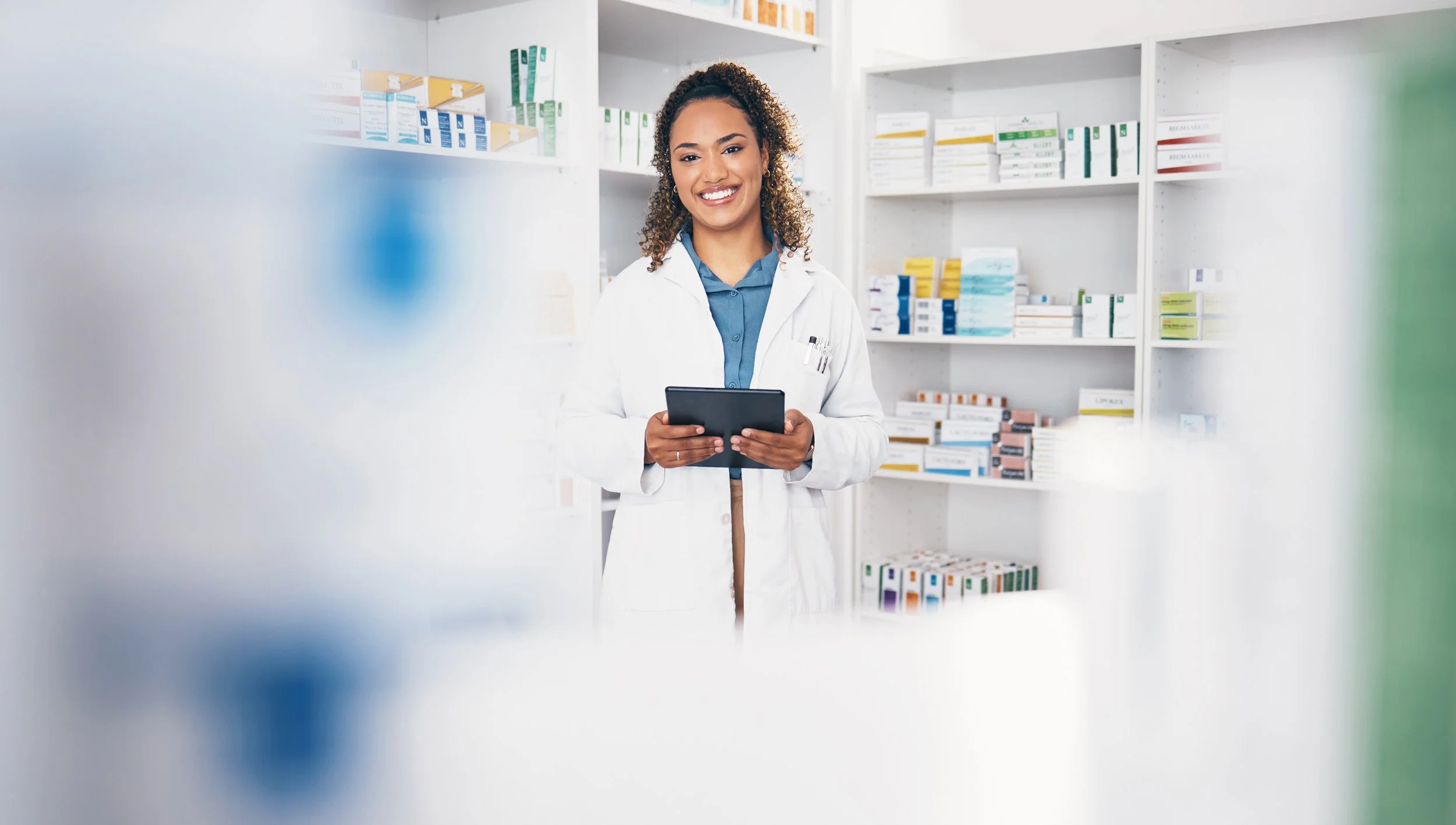 A smiling female pharmacist standing in a pharmacy, holding a black tablet device, with shelves of medication boxes in the background.