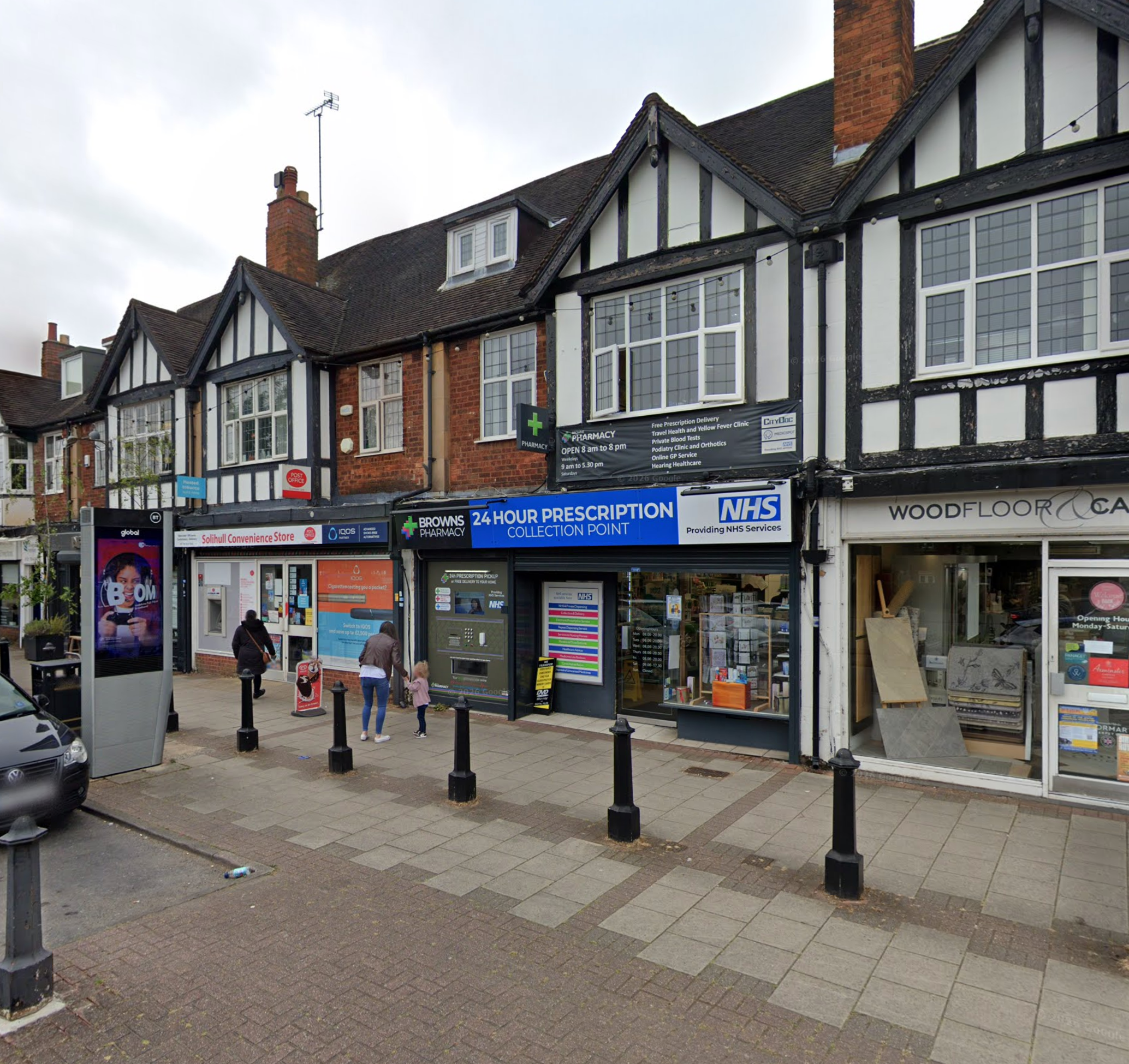 Street view of a shopping area with shops, people walking, and a cloudy sky. The shops include a pharmacy, a convenience store, and other businesses. There are black bollards on the sidewalk and a few cars parked nearby.