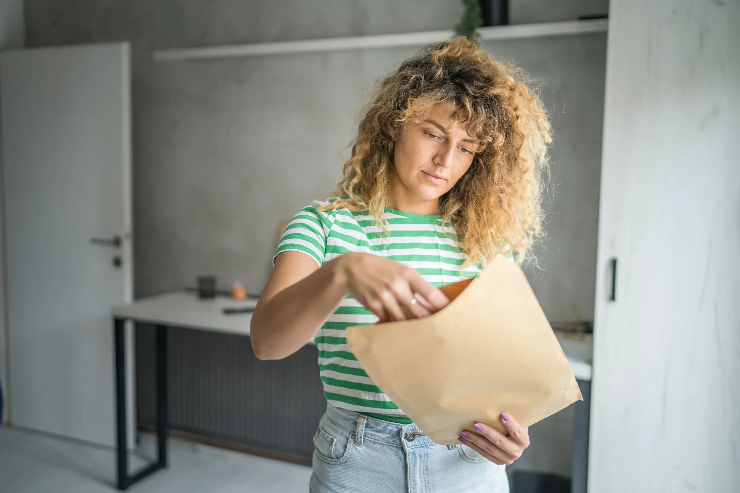 A young woman with curly blonde hair wearing a green and white striped shirt and light blue jeans is opening a brown paper bag in a modern, minimalist room with concrete walls and a black desk.