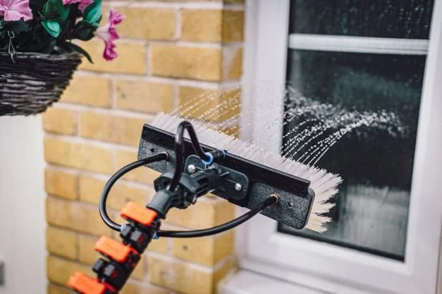 A pressure washer with a scrubbing attachment cleaning a window outside a brick house.