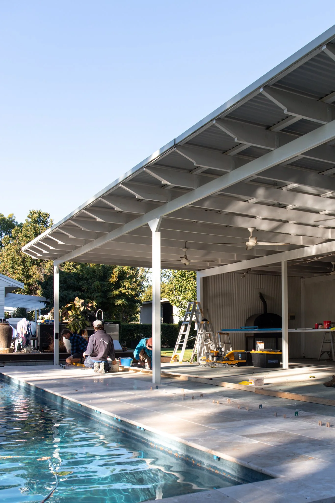 People working under a large metal roof structure next to a swimming pool, with tools and ladders around them, in a backyard setting.