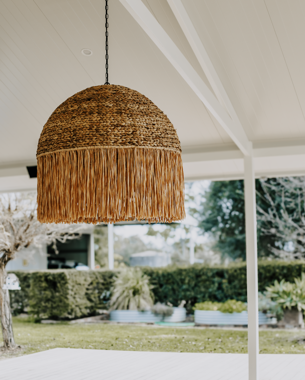 A woven rattan pendant light with frayed edges hanging from a chain outside a porch area with greenery in the background.