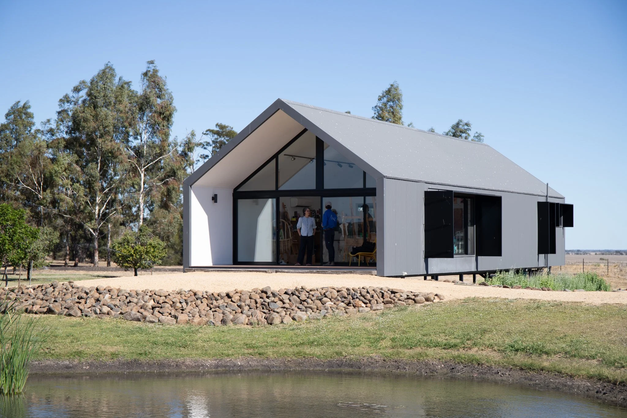 Modern house with large windows and open garage, situated near a pond and surrounded by trees and grass under a clear blue sky.