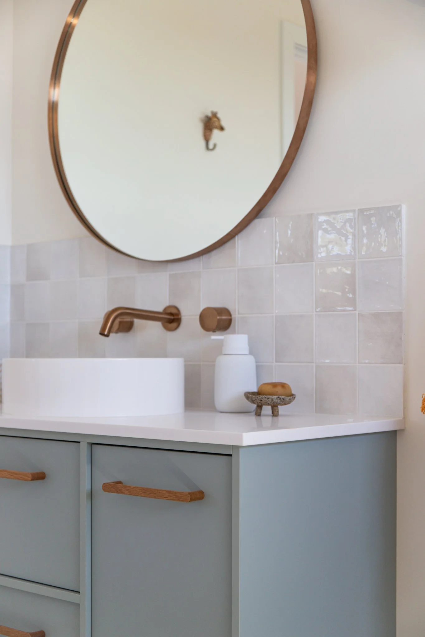 A bathroom vanity with a light blue cabinet, wooden drawer handles, a white countertop, a gold faucet, a white soap dispenser, a small decorative stone dish with a bar of soap, a round mirror with a wooden frame, and a wall-mounted gold hook in the shape of an animal head reflected in the mirror.