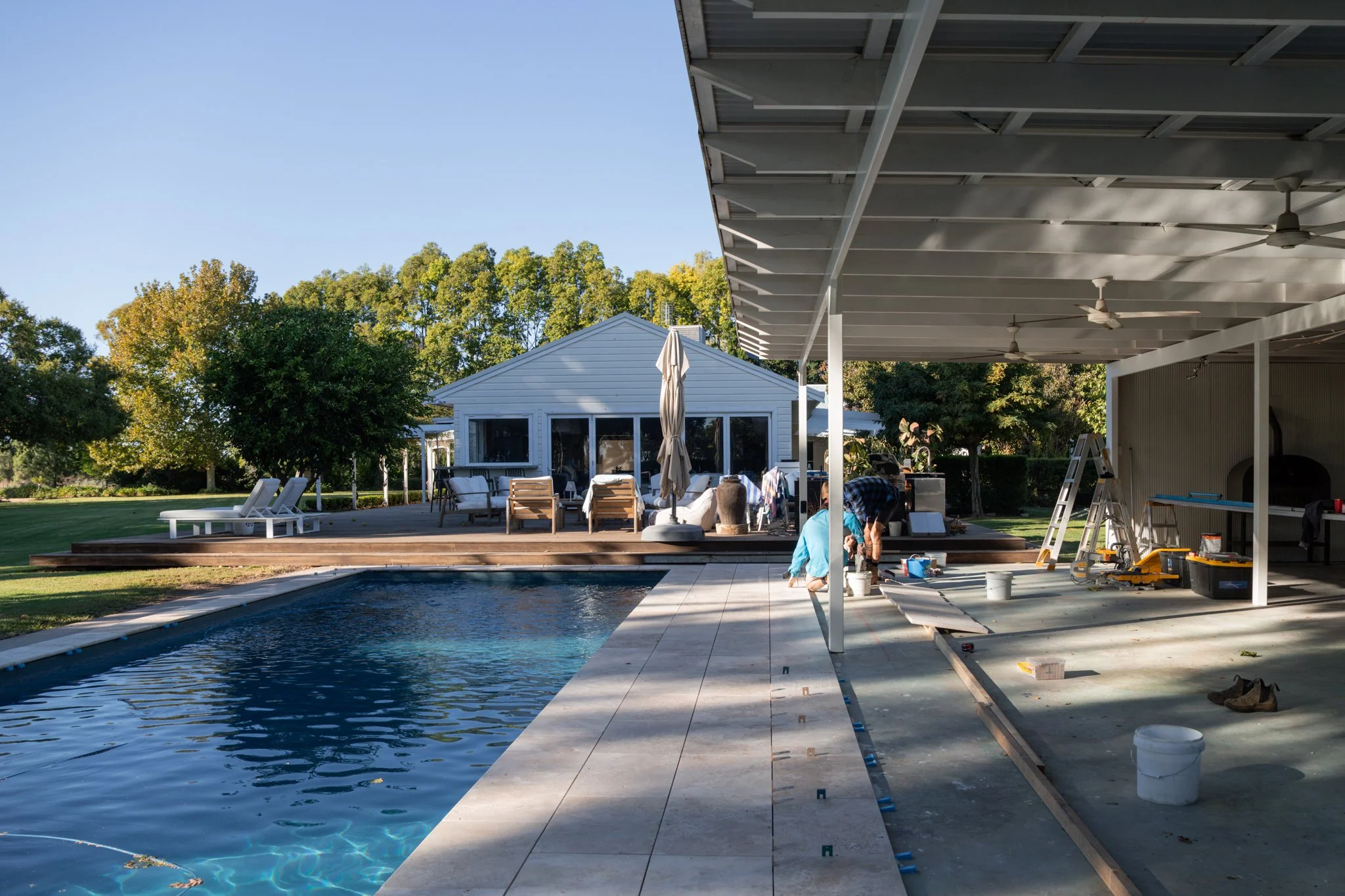 Backyard with swimming pool, patio furniture, and house under construction or renovation, with workers and tools.