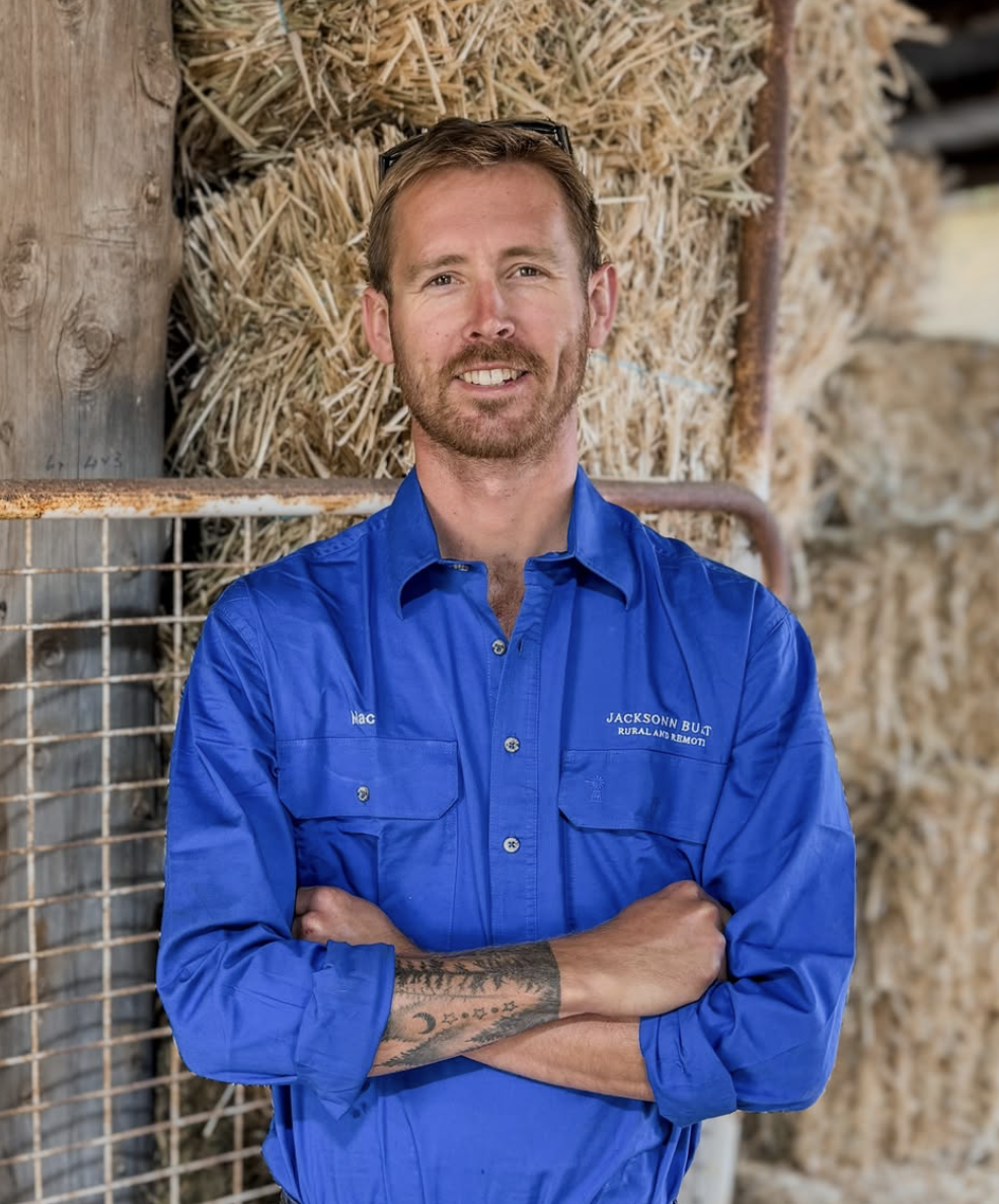 A man with light skin, wearing a blue shirt, standing with arms crossed in front of a stack of hay bales in a barn or farm setting.