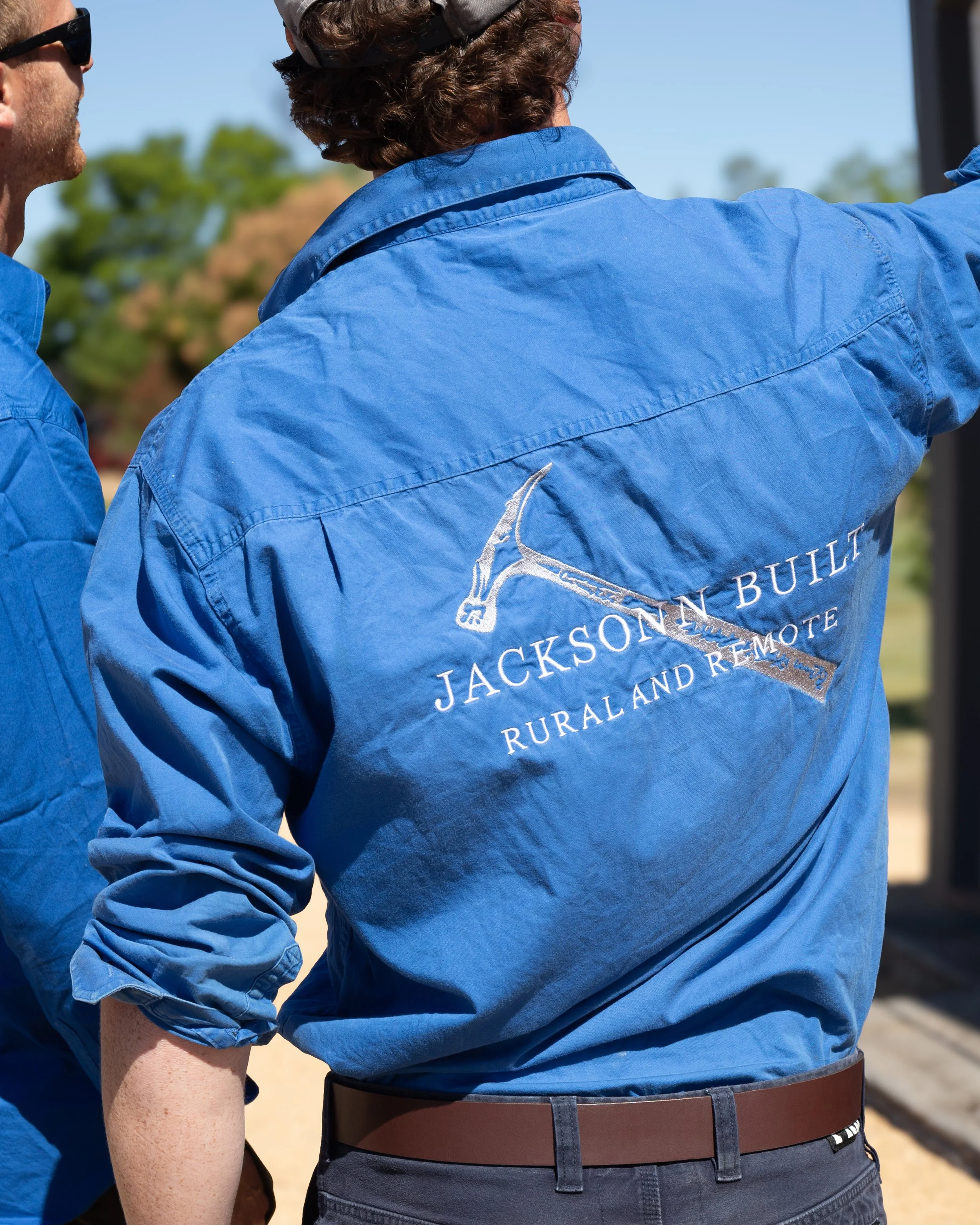 Close-up of two men in blue shirts, one with rolled-up sleeve, outside with blurred trees in background.