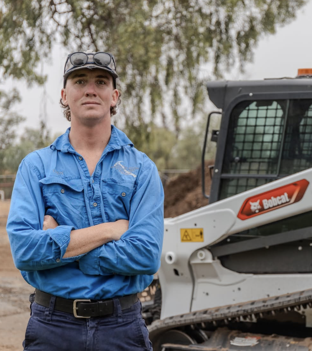 A man with arms crossed wearing a blue work shirt, standing outside in front of a Bobcat skid-steer loader.