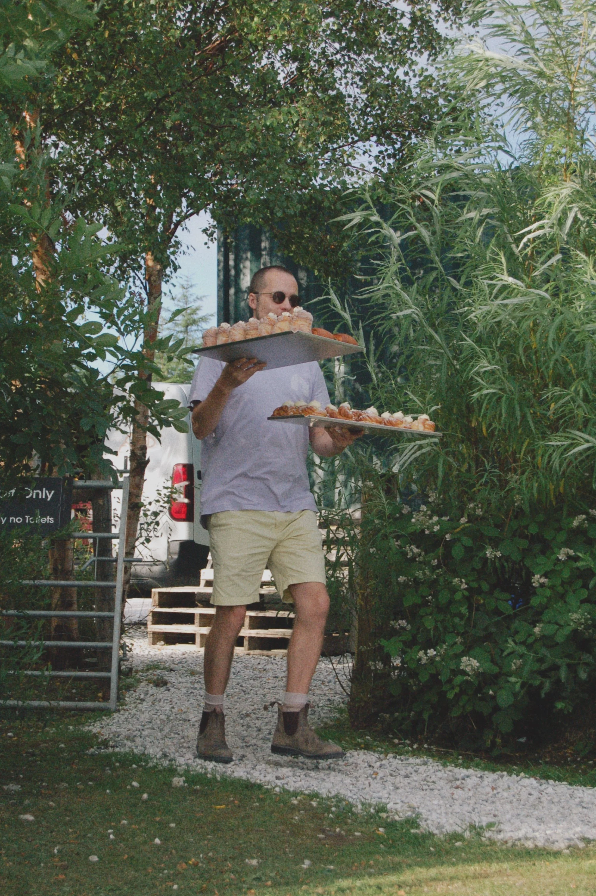 A man wearing sunglasses, light-colored shorts, and boots carries trays of food outdoors surrounded by greenery.