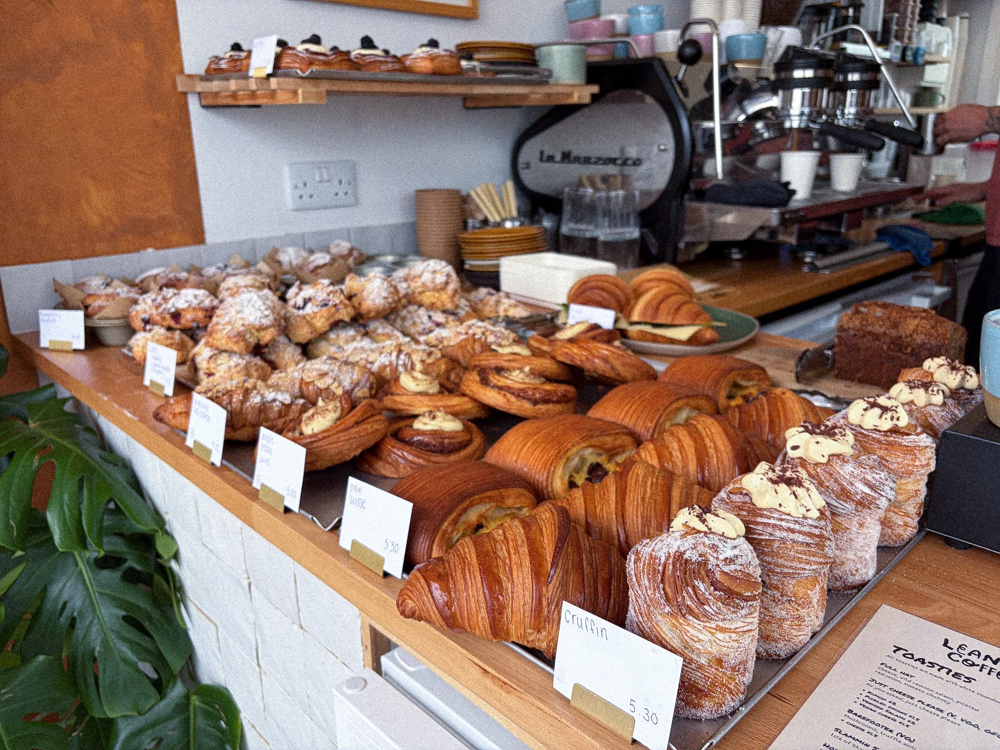 Display of assorted baked goods, including croissants, danishes, and pastries, on a bakery counter. There are small signs with prices and labels like "Croissant" and "Danish" in front of the items.
