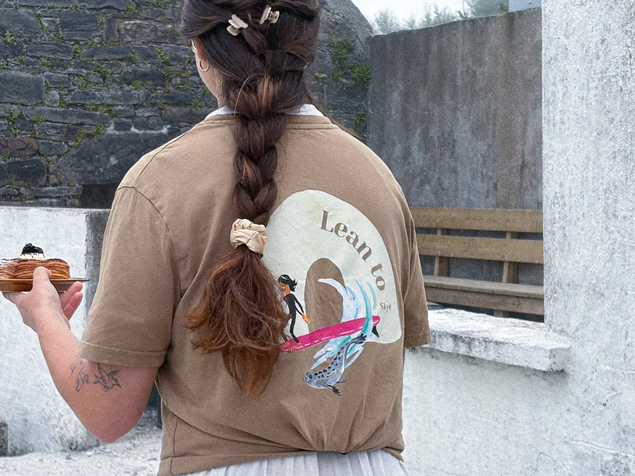 Person with long braided hair holding a plate of food. The person is wearing a beige t-shirt with a dolphin graphic and the words 'Learn to' visible. An outdoor stone wall is in the background.