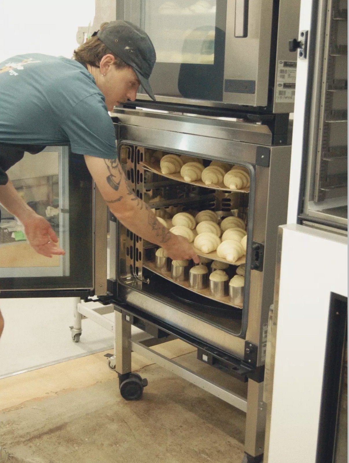 A person with tattoos and a cap putting croissants into a commercial oven with multiple shelves.