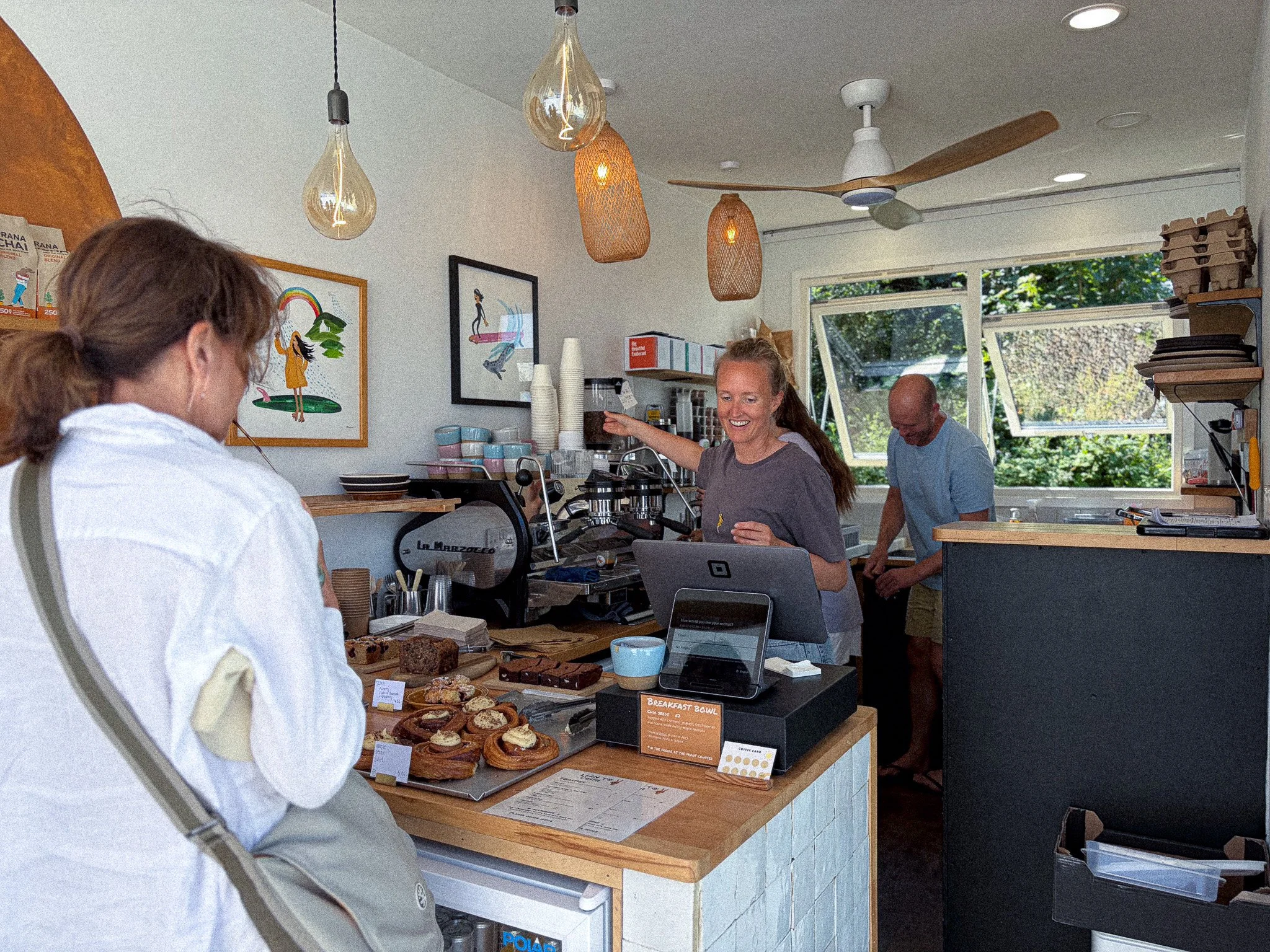 A coffee shop or bakery with people preparing and serving baked goods, with hanging light fixtures and framed artwork on the wall.