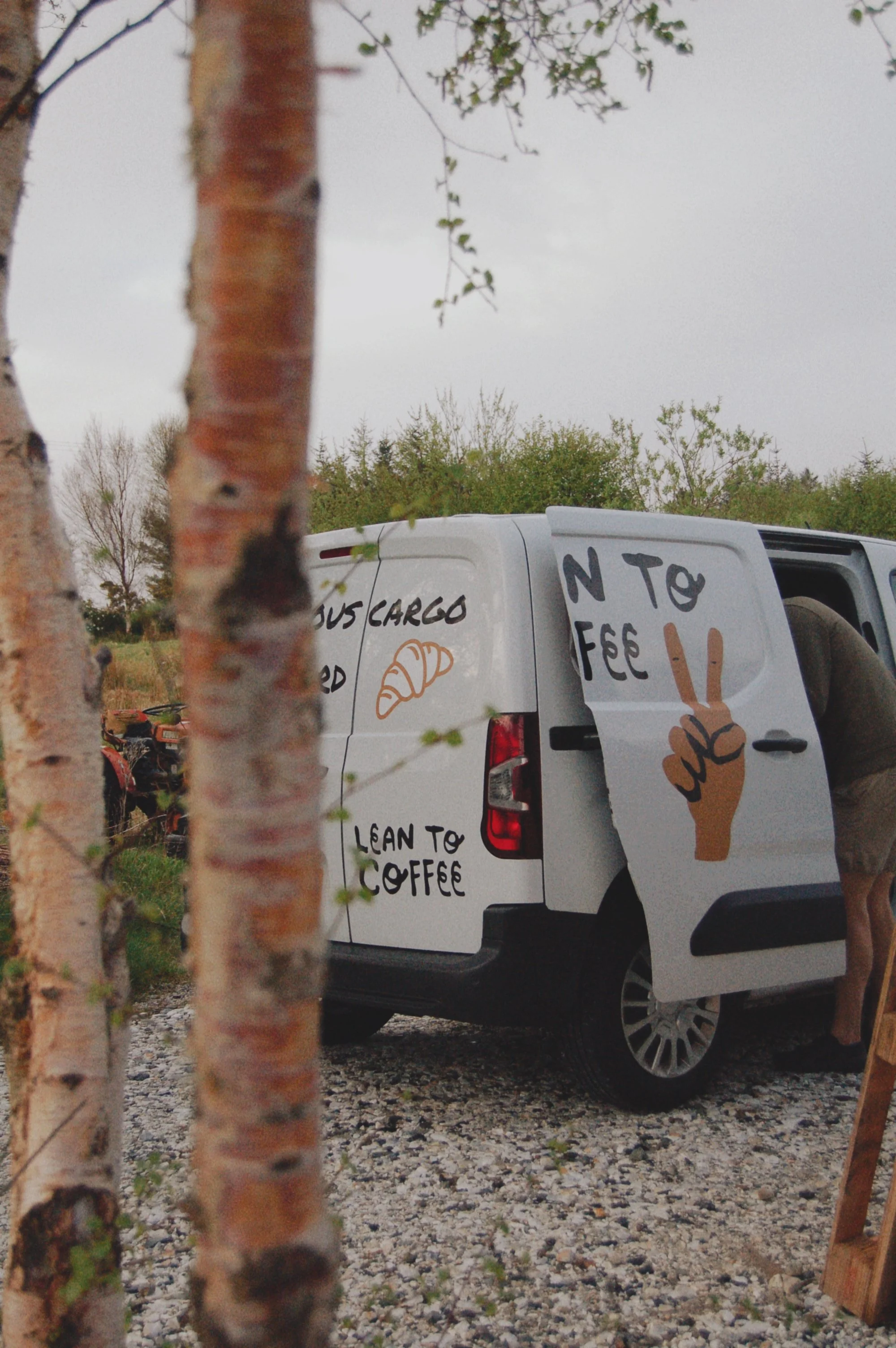 A white van with a sign on the back door that features a cartoon hand making a peace sign and the words "N to F to Coffee," parked outdoors on a gravel surface. There are trees in the background and some branches in the foreground.