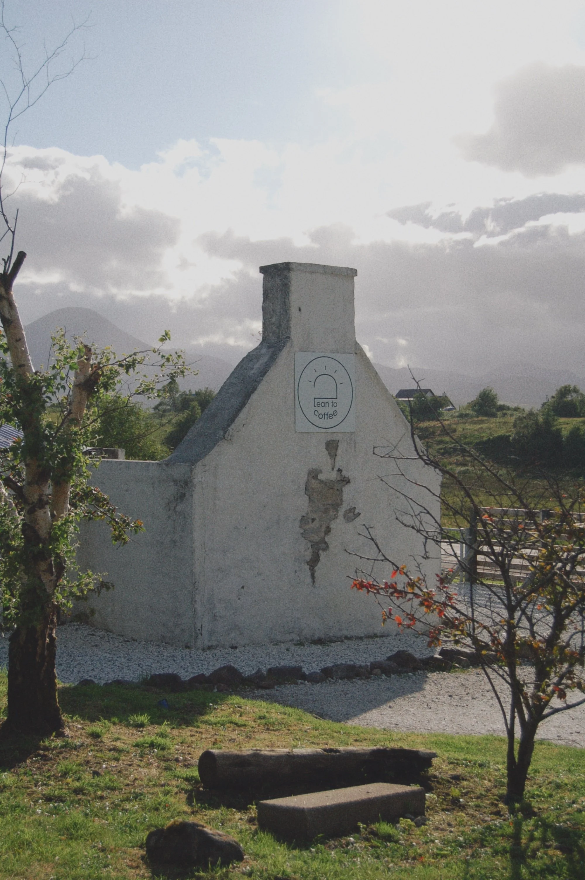 A white wall with a sign that reads 'Lean to Coffee' hangs beneath a small chimney. The wall shows some cracks. There are trees around, a grassy area, mountains and cloudy sky in the background.