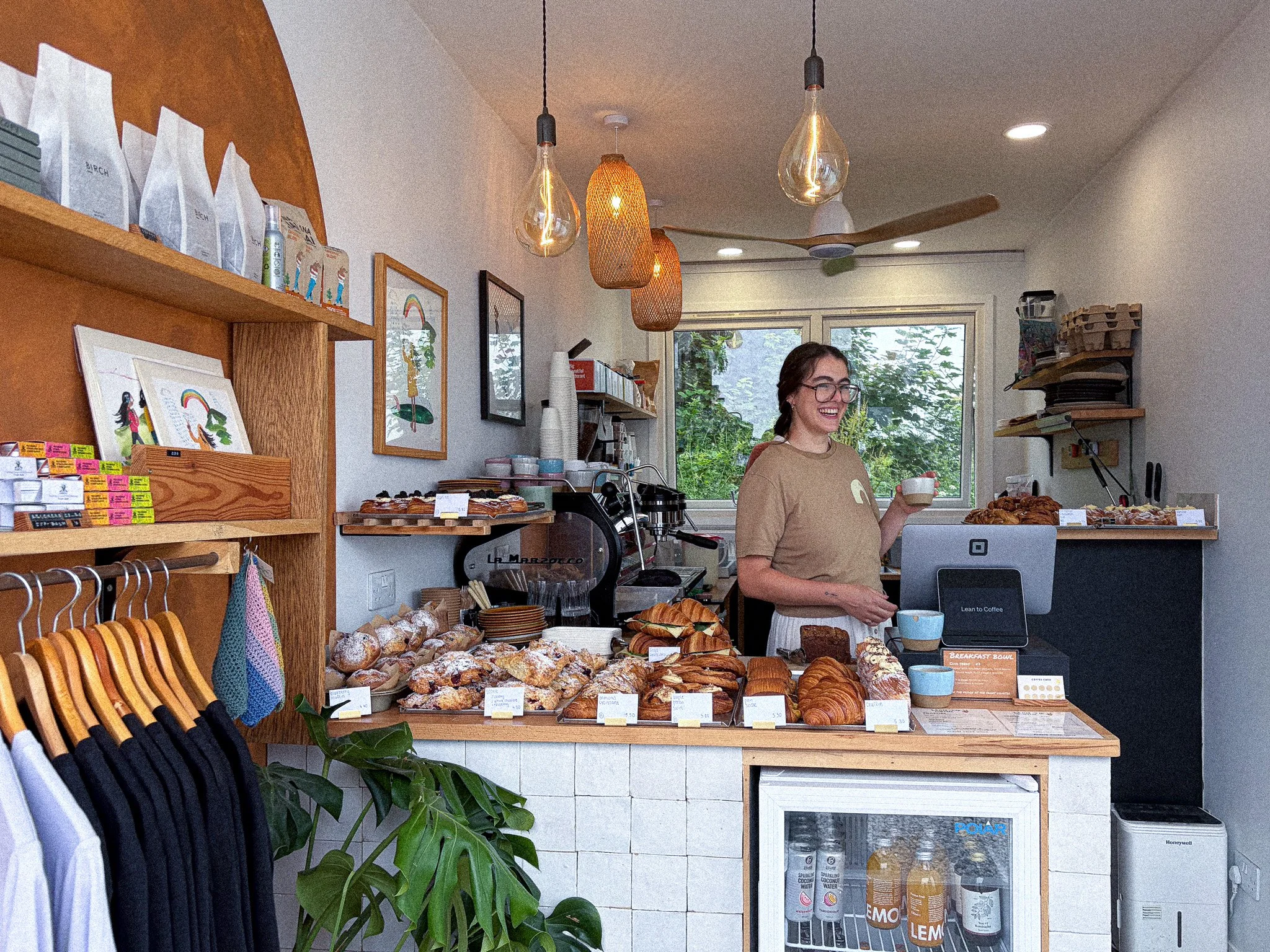 A woman with glasses and a beige shirt stands behind a counter with various baked goods such as croissants and muffins. The shop interior features wooden shelves with products and artwork on the walls, pendant lights hanging from the ceiling, and a large window showing greenery outside.