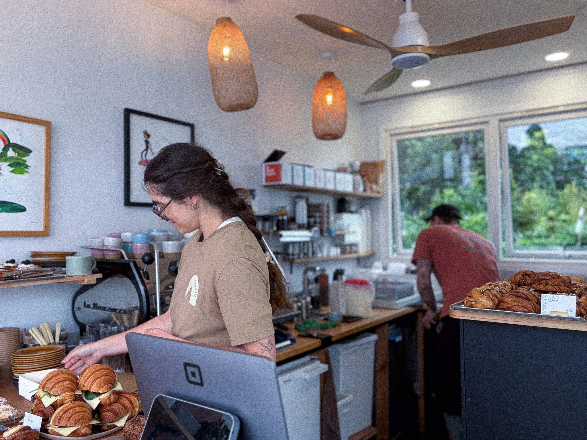 A woman with dark hair and glasses preparing baked goods in a bakery or cafe. There are croissants and other pastries on the counter, and a man is sitting in the background near a window.