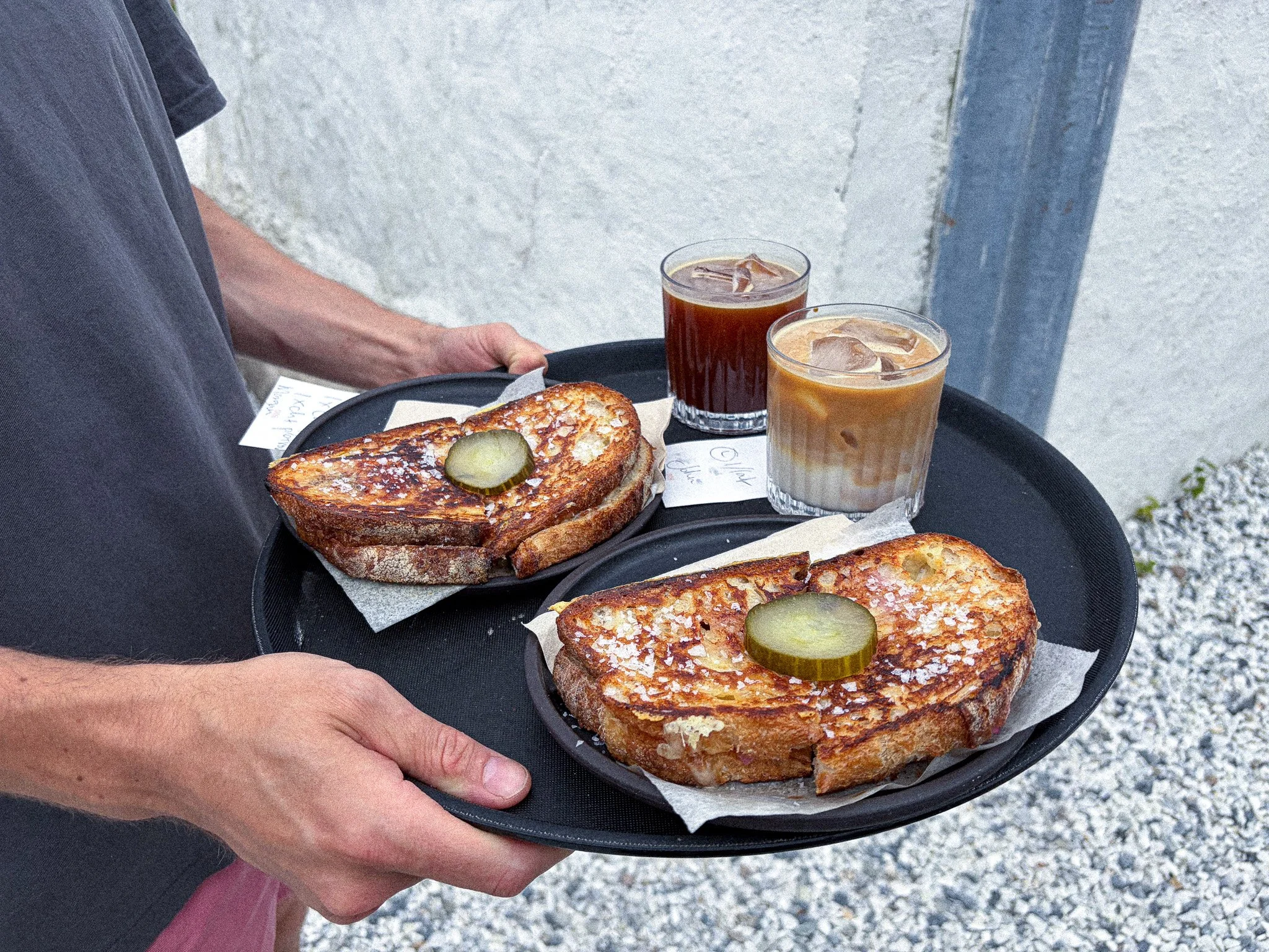 Person holding a tray with toasted sandwiches topped with pickles, two glasses of iced coffee, and a piece of paper on a black tray, against a textured gray wall background.