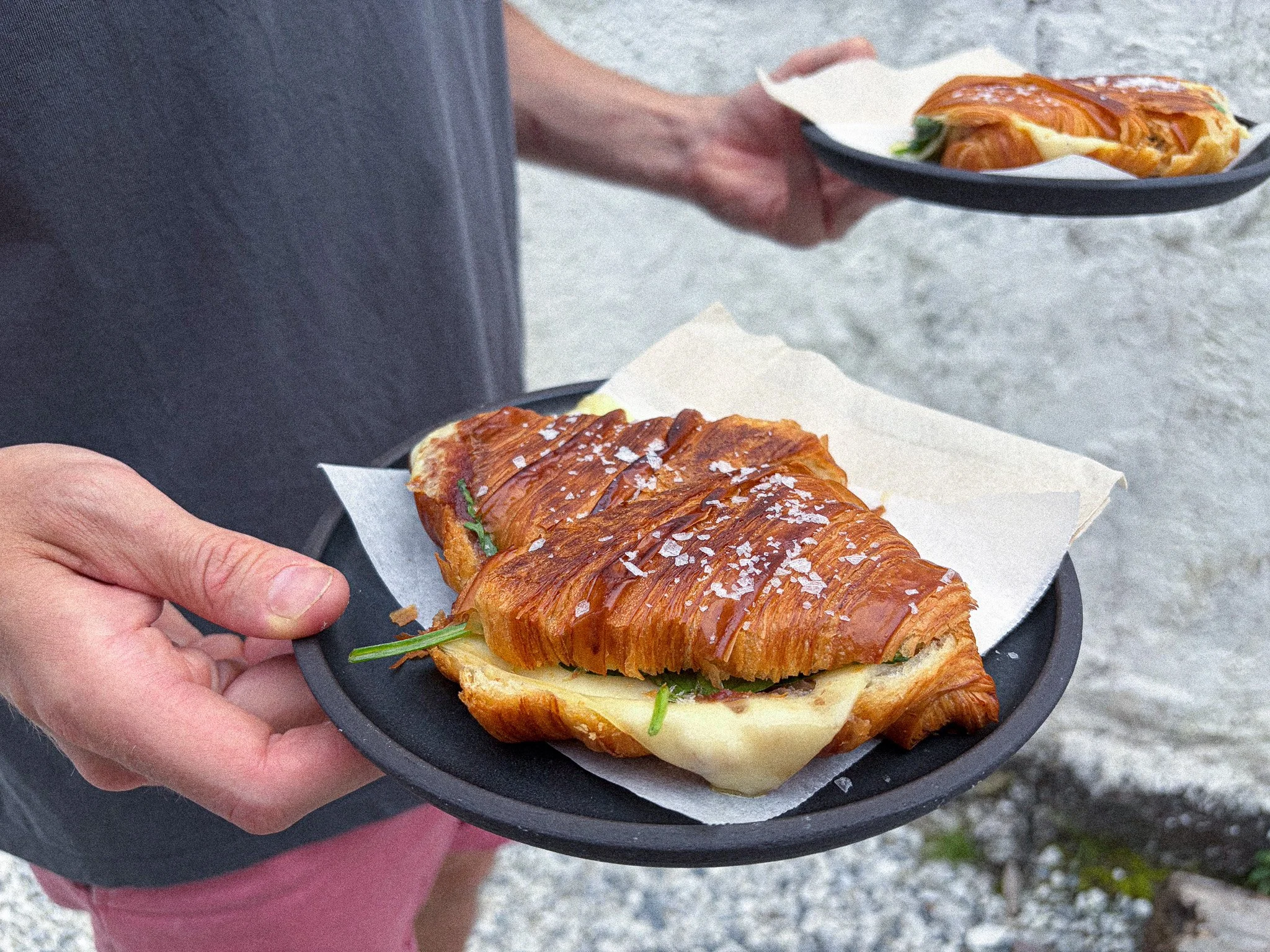 A person holding a small black plate with a sandwich featuring a croissant bun, cheese, lettuce, and a meat filling, with coarse salt sprinkled on top, outdoors.