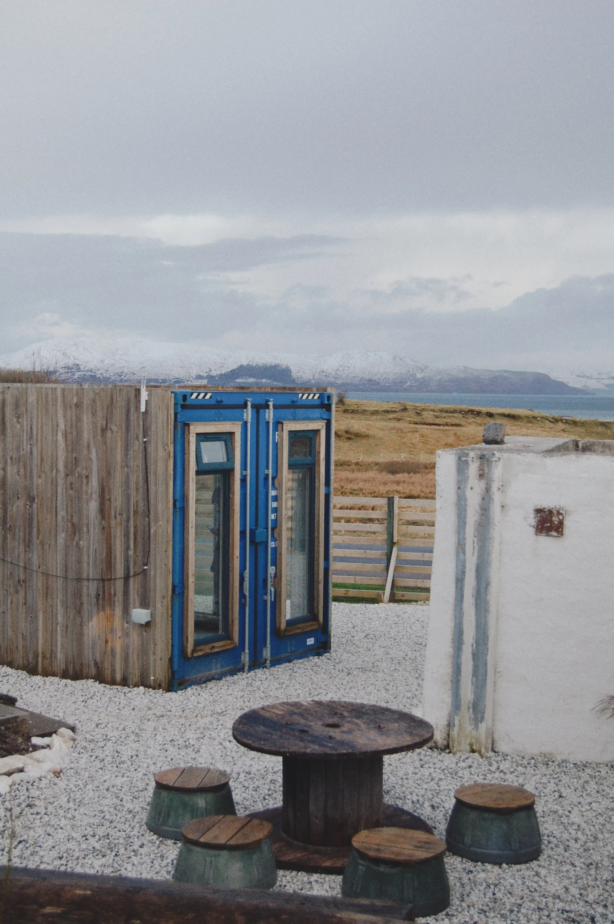 An outdoor scene with a round wooden table and four matching stools with green bases on gravel, a small white wall, a blue phone booth, a grassy field, and snow-capped mountains in the background under a cloudy sky.