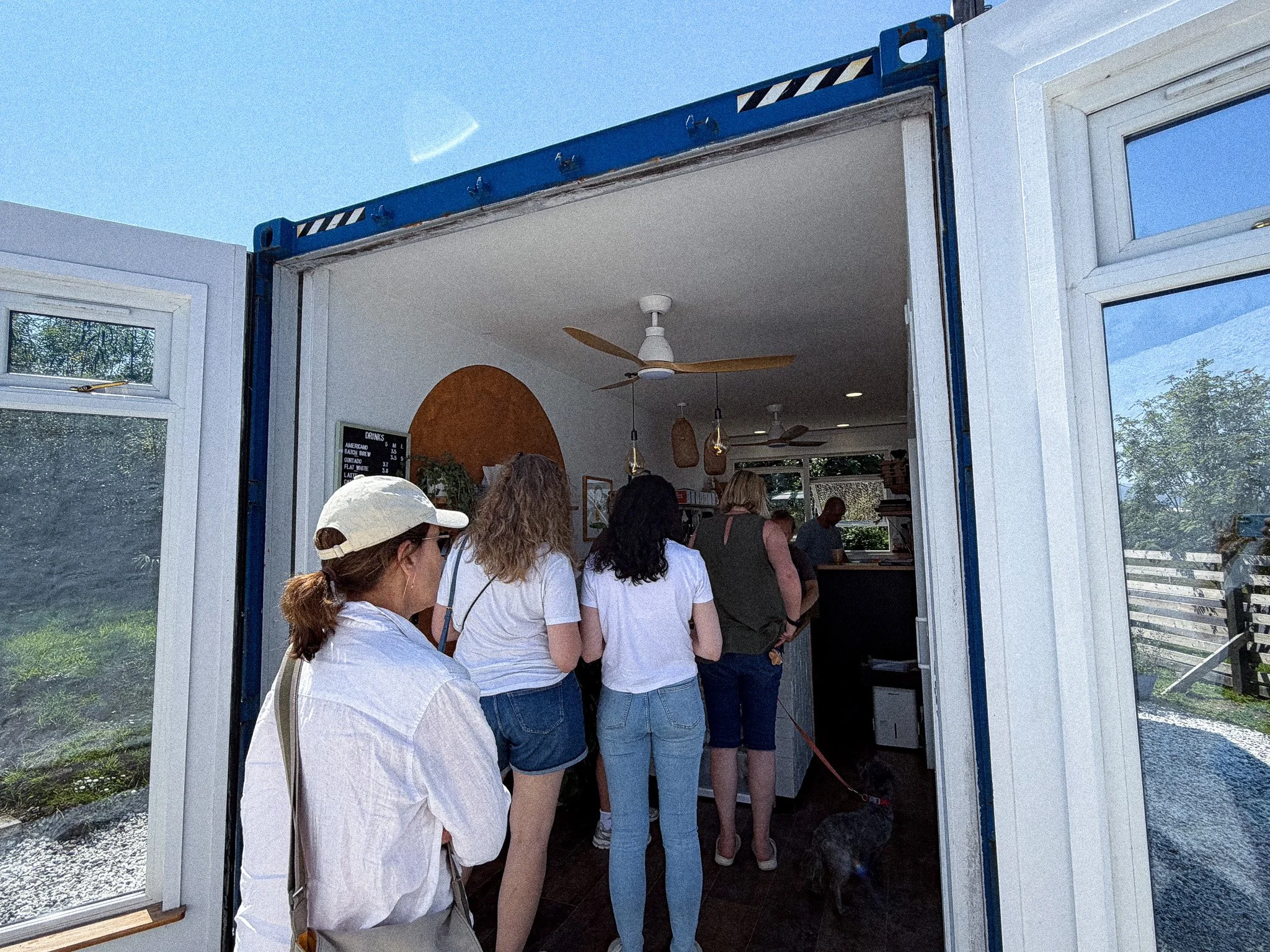People standing in line inside a cafe or bakery, viewed from outside through a doorway, with a blue sky visible above.