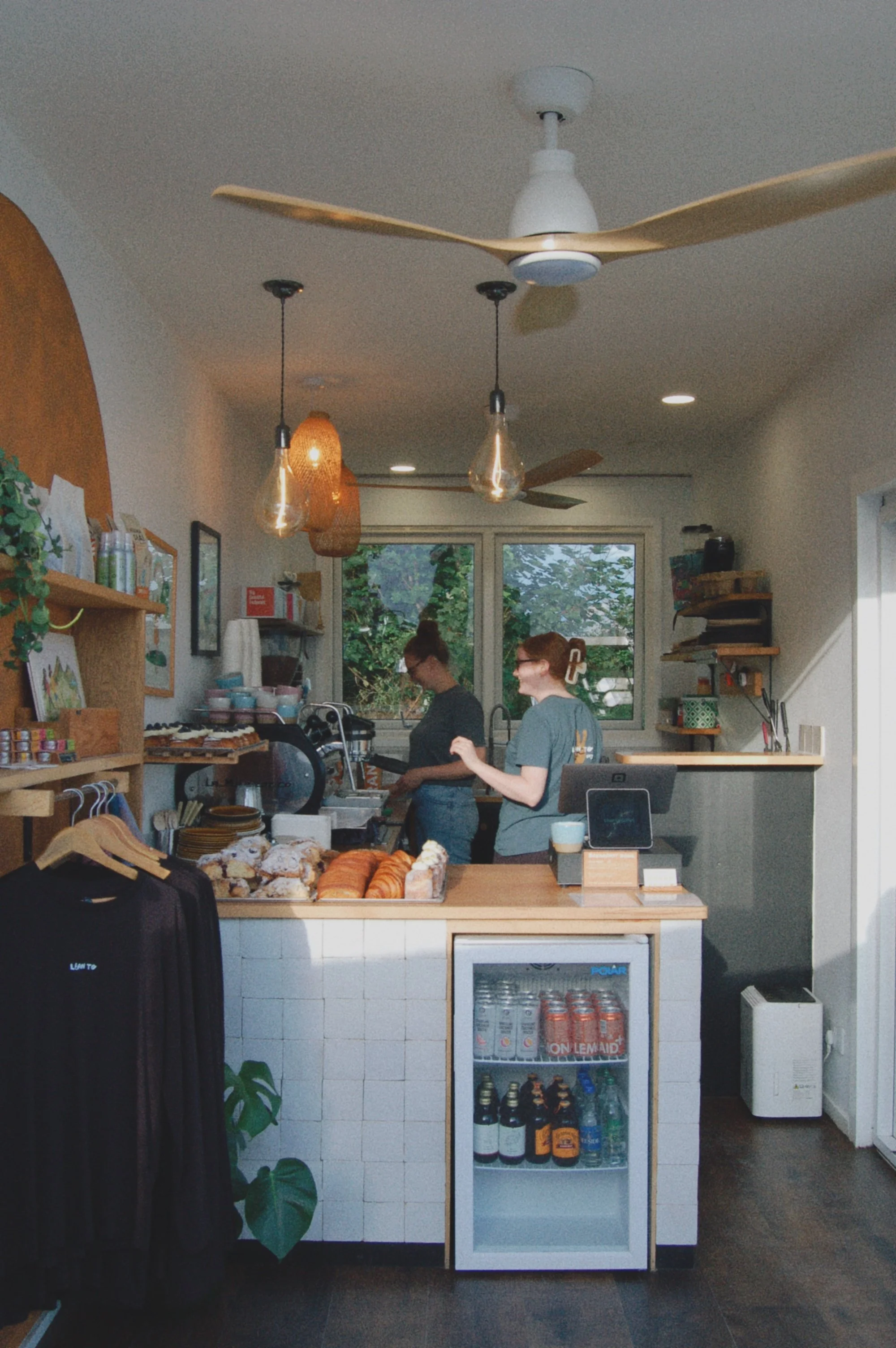 Inside a cozy cafe or bakery with baked goods on display, two women working behind the counter, warm lighting, and a window showing greenery outside.