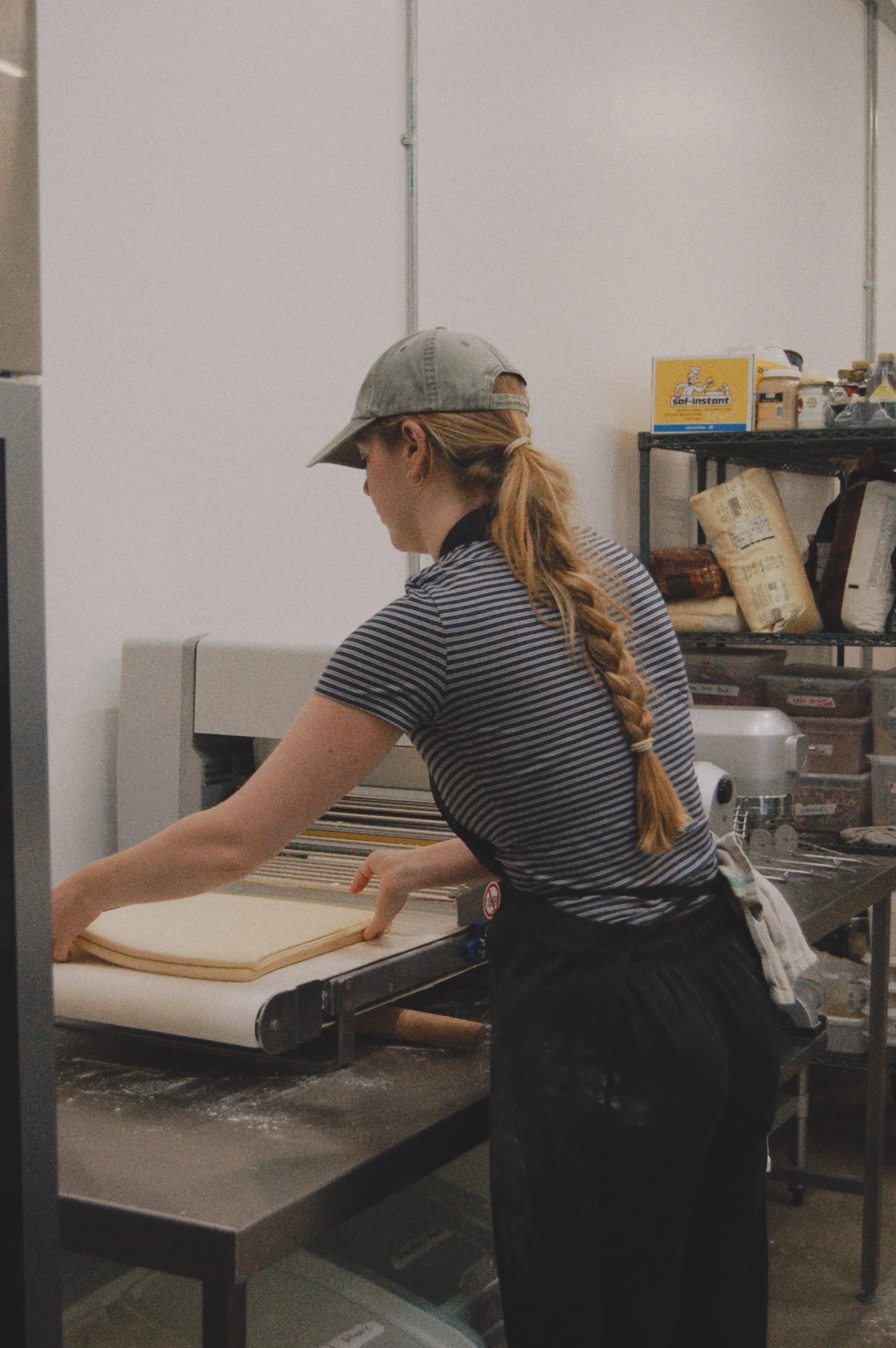 A woman with a long braid wearing a baseball cap and striped shirt presses dough flat on a work table in a commercial kitchen or bakery, with shelves of supplies in the background.