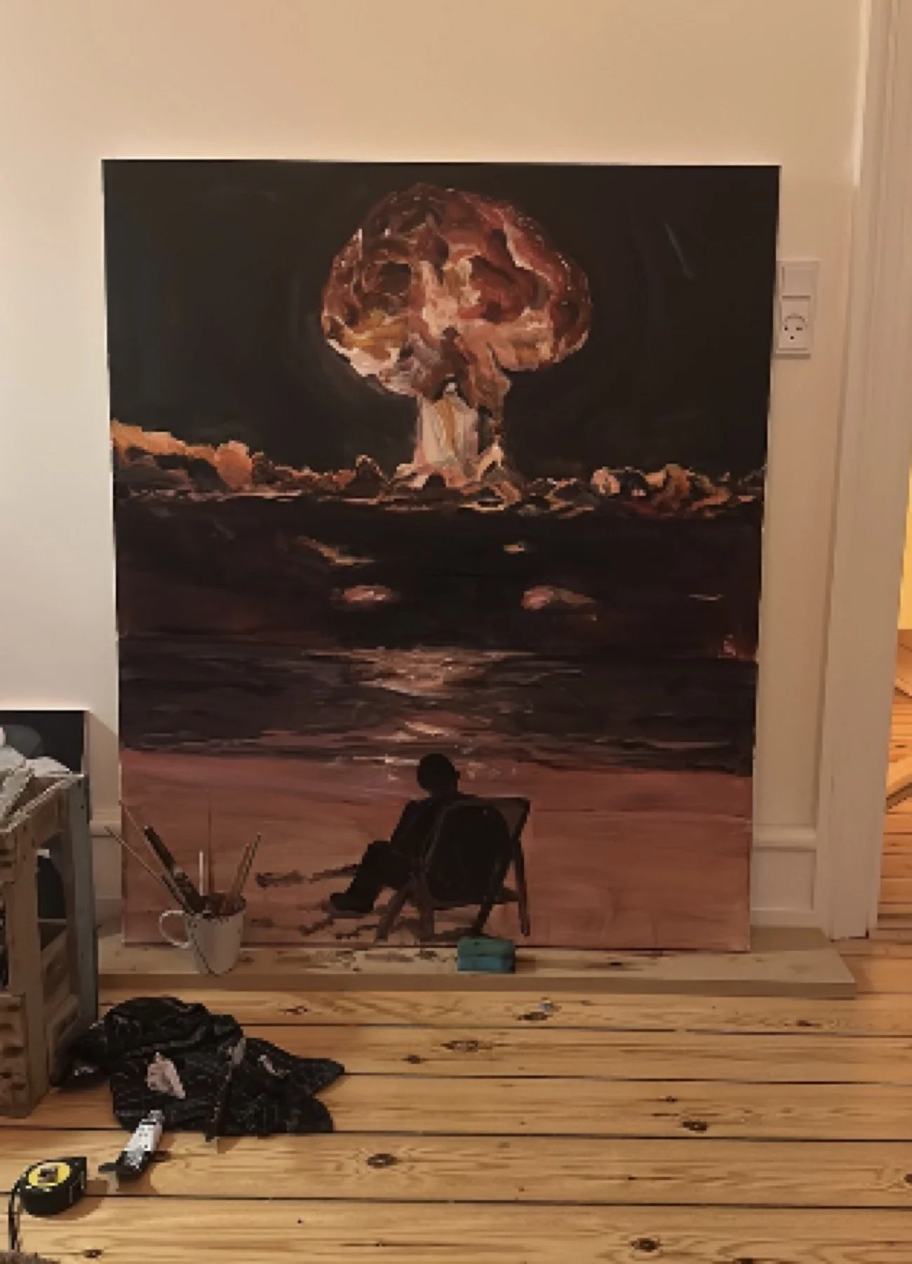 Et værelse med et stort kunstværk foran en væg, der forestiller en eksplosion eller en atombombe. En person sidder i en stol ved væggen, og der er værktøj og materialer spredt på gulvet.