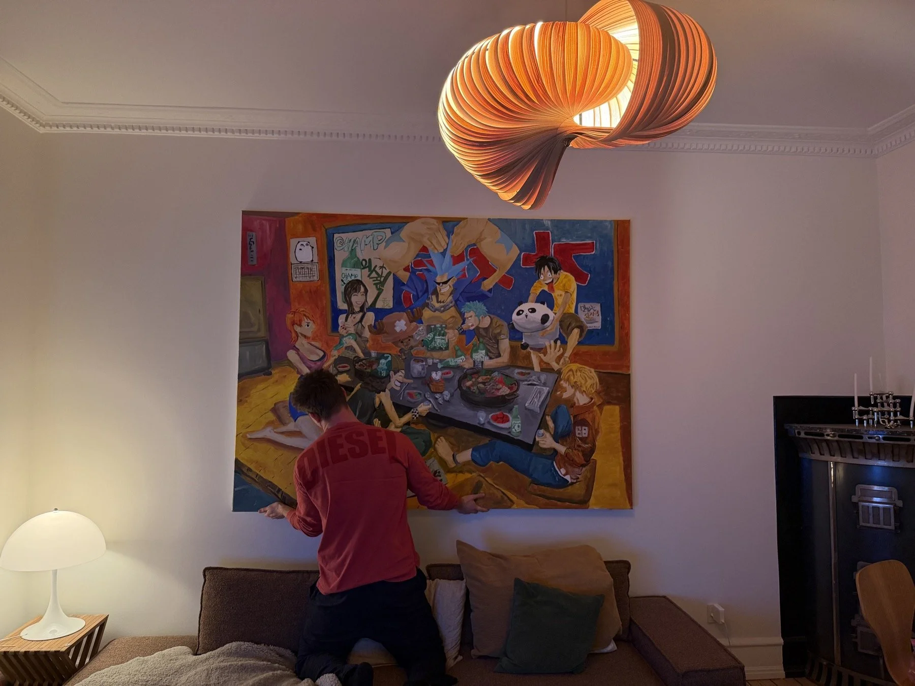 To personer står foran et stort farverigt maleri på væggen i en stue. En af dem har rød sweater og sæder på gulvet med puder, og der er en lampe på bord ved siden af.