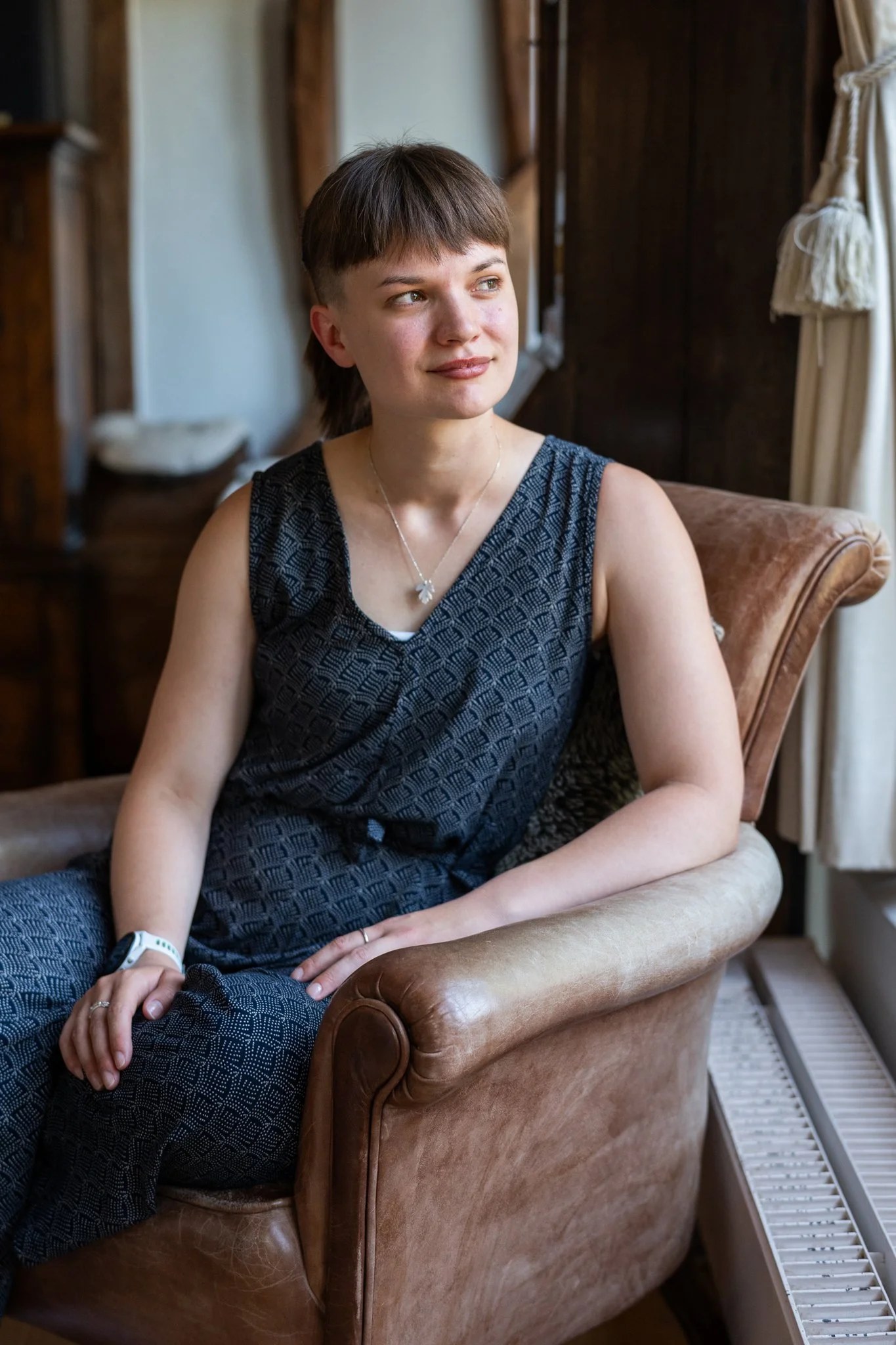 A young woman sitting on a leather armchair indoors, with her hands resting on her lap, looking to the side with a slight smile. She has short brown hair and is wearing a dark patterned sleeveless top, a necklace, and a white wristband. The background includes wooden furniture and light-colored curtains.