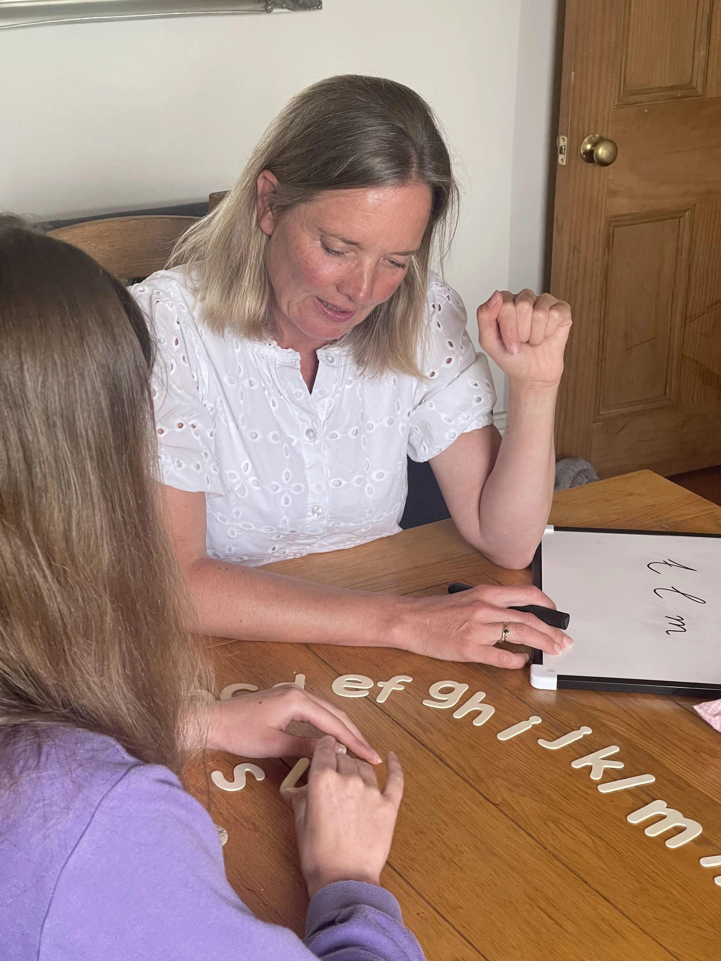 Two women sitting at a wooden table, spelling out words with white alphabet letters on the table, and one woman writing on a whiteboard.