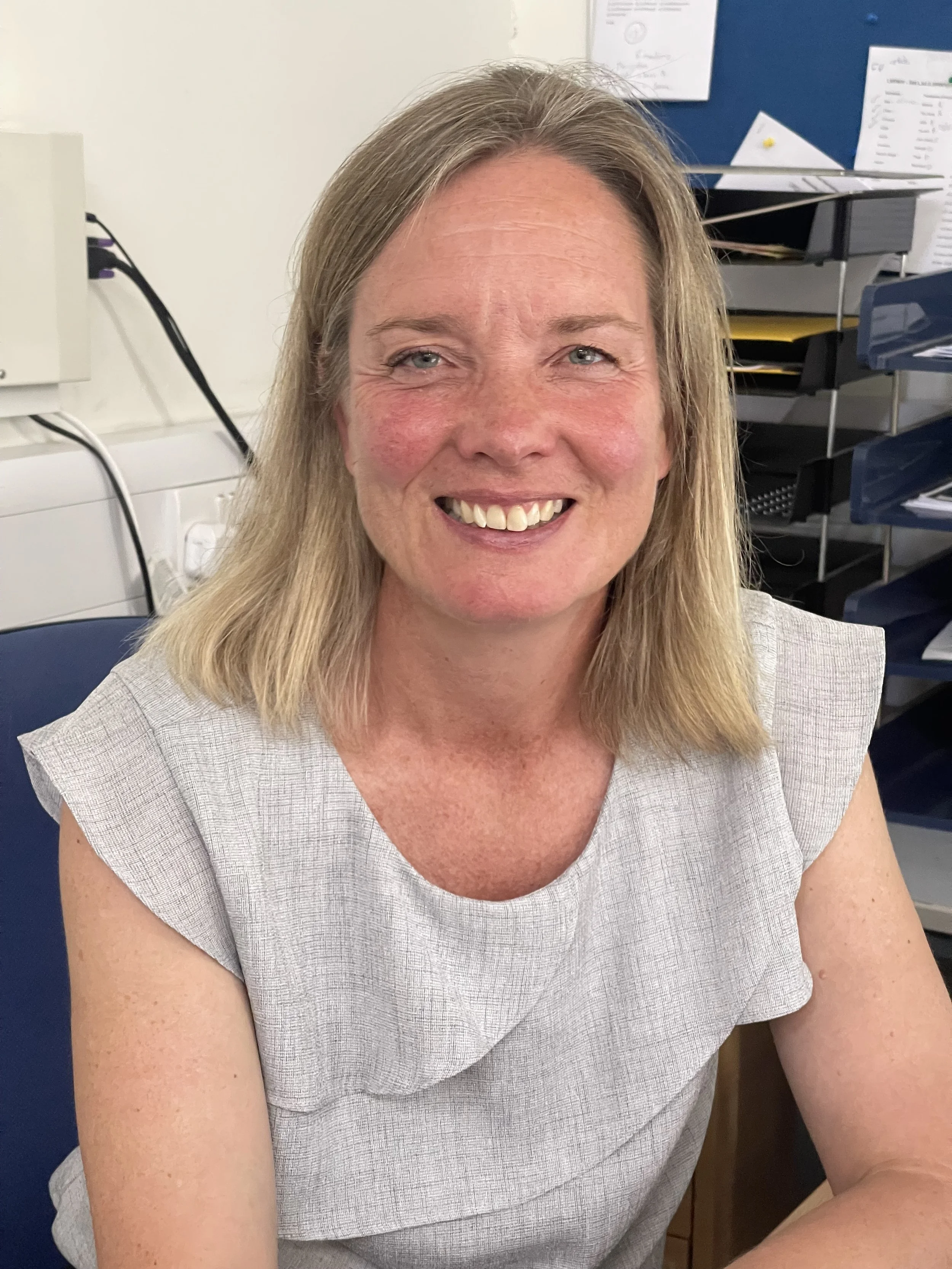 Close-up of a middle-aged woman with blonde hair smiling, wearing a light gray top, in an office setting with papers and shelves in the background.