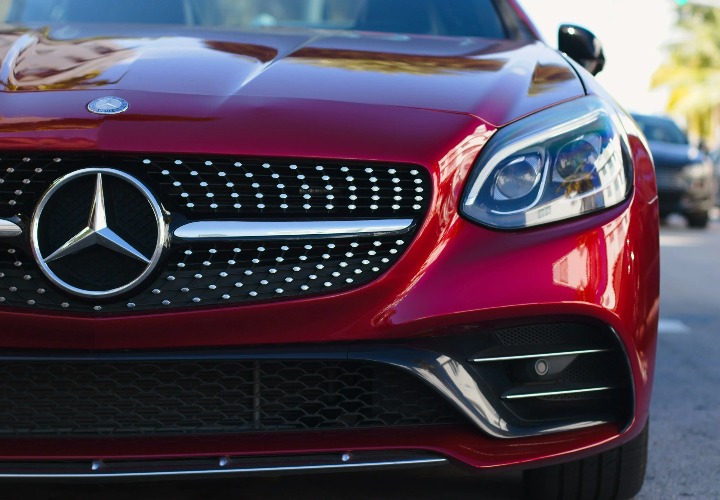 Red Mercedes-Benz car front close-up, showing grille, logo, and headlight, parked outdoors.
