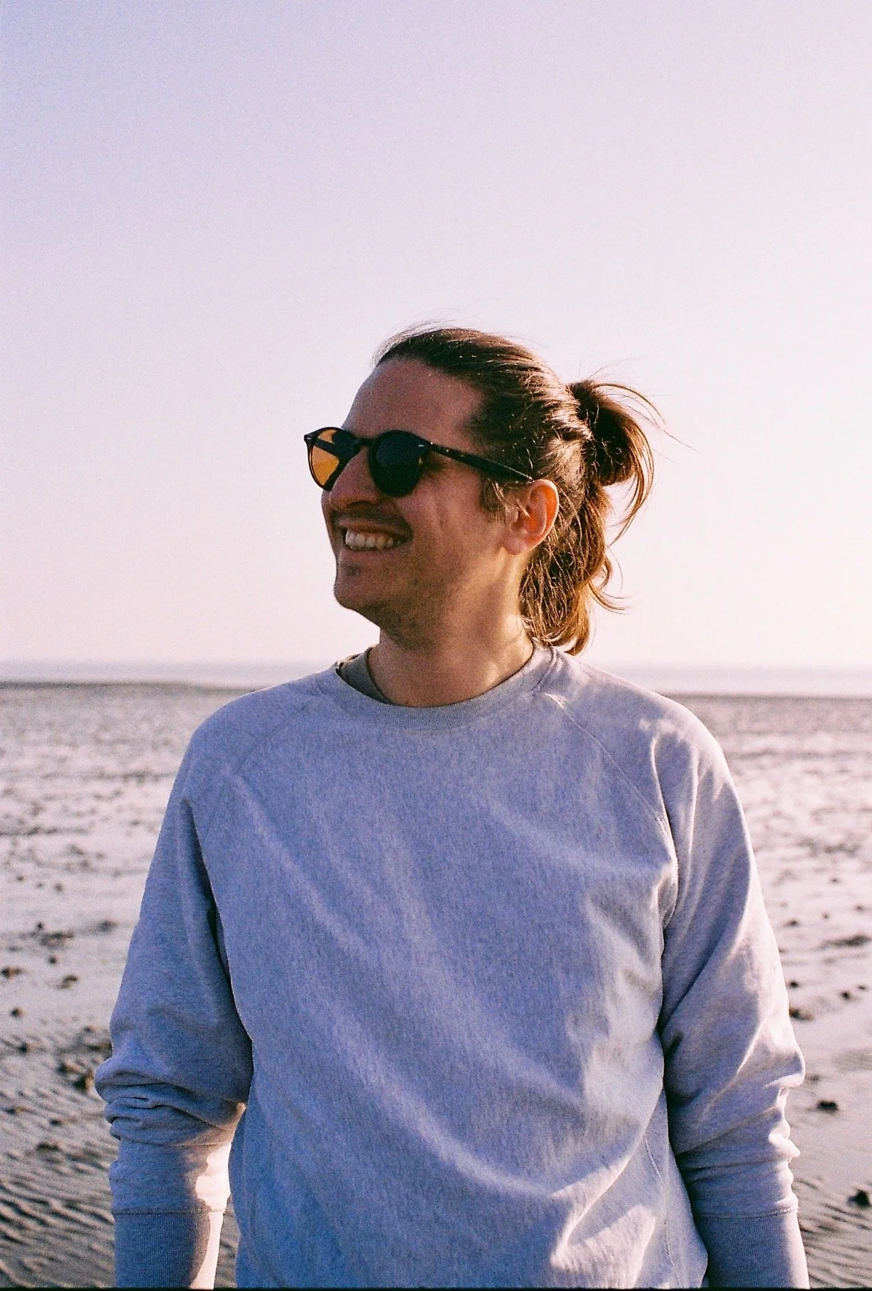 Eine lachende Frau trägt Sonnenbrille und einen grauen Pullover am Strand bei Sonnenuntergang, im Hintergrund Meer und Sand.