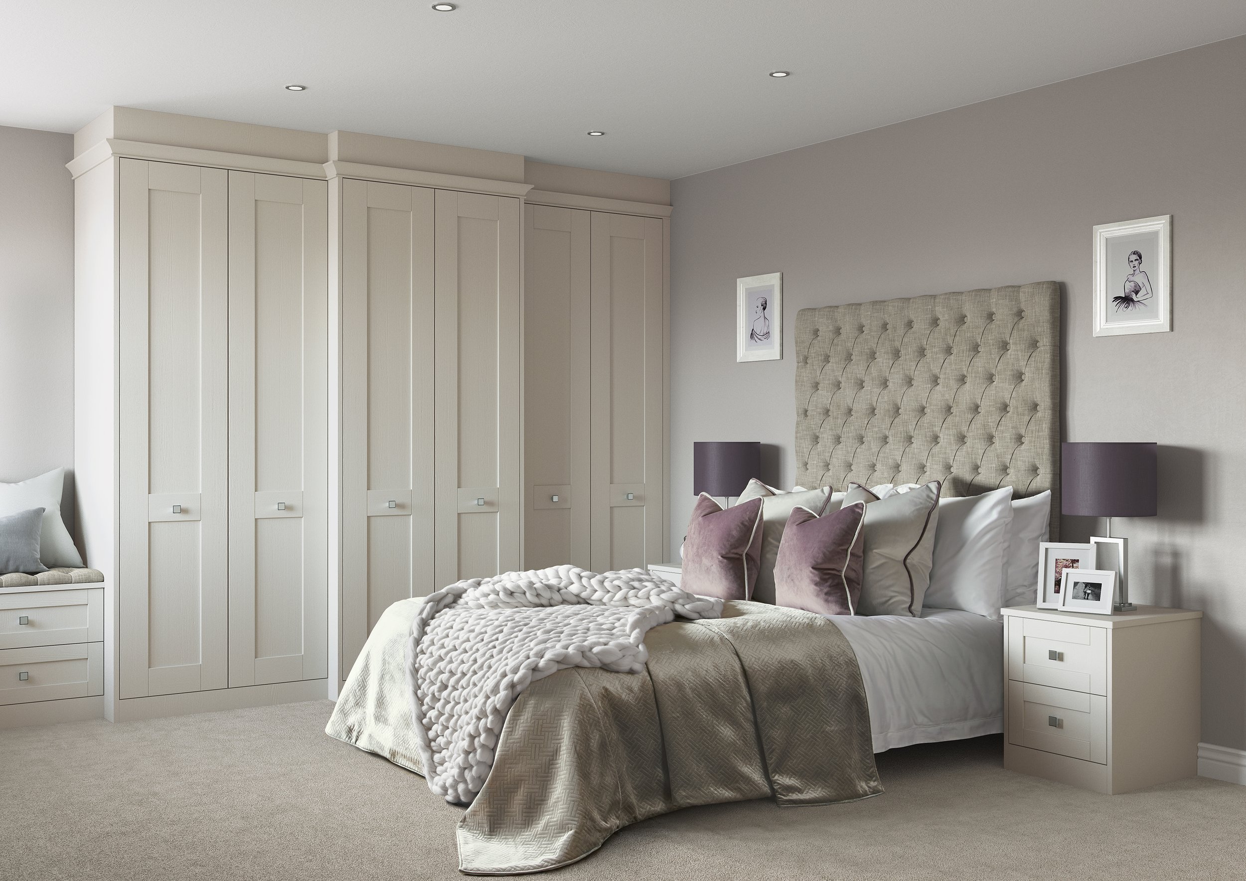 A bedroom with a large bed featuring an upholstered headboard, purple pillows, and a chunky knit throw blanket. There are two bedside tables with lamps, framed pictures, and a small sitting area with cushioned seating. The room has neutral, light-colored walls, a built-in wardrobe, and recessed ceiling lights.