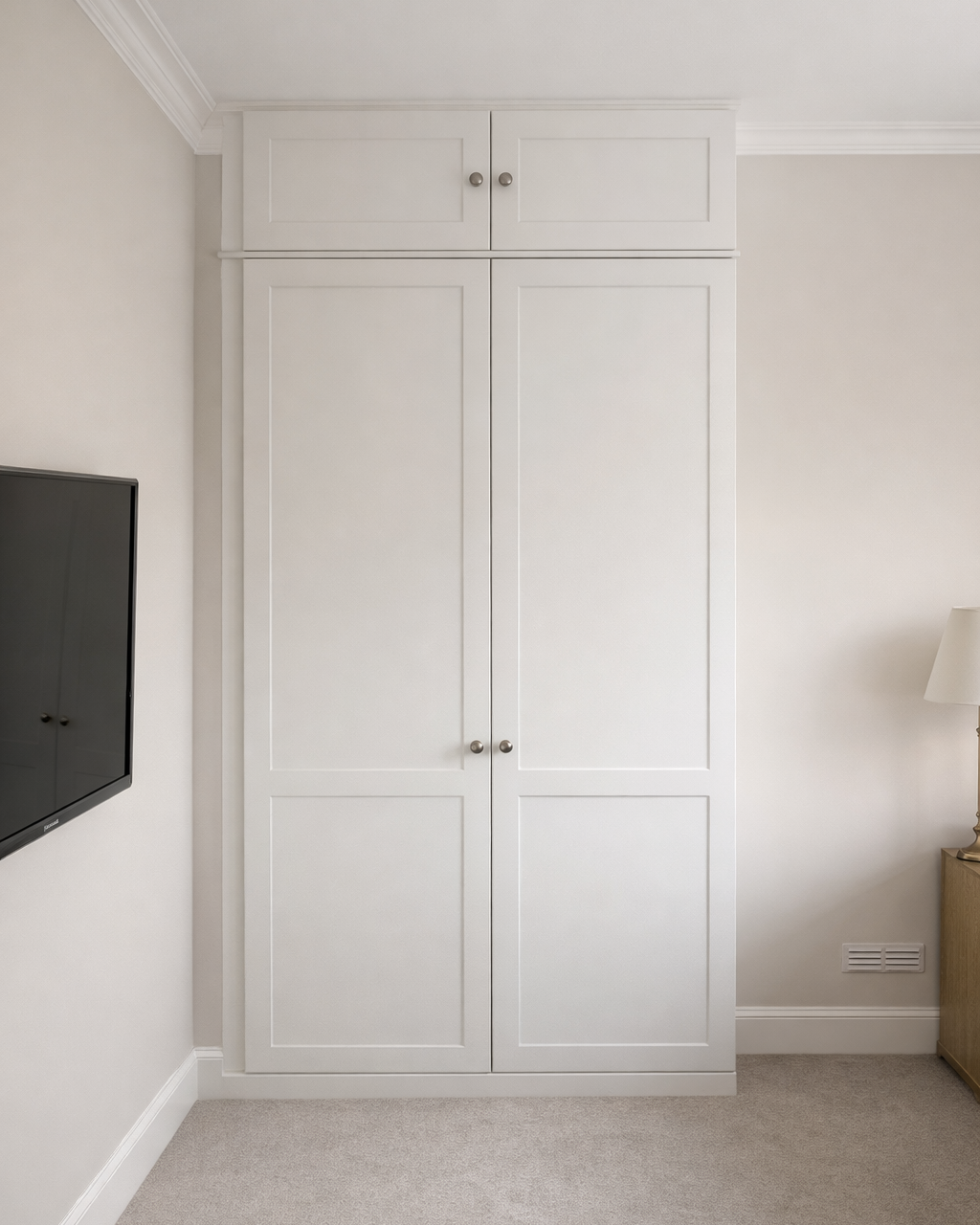 A tall, white built-in closet with two doors in a room with light-colored walls, a flat-screen TV mounted on the wall, and a wooden nightstand with a lamp on top.