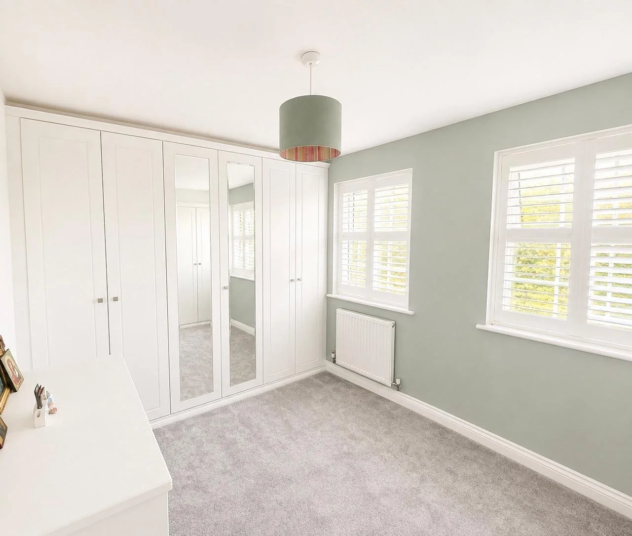 Bright bedroom with white built-in wardrobes, two large windows with white shutters, a radiator below the windows, a white dresser with photo frames and small items, and a ceiling light fixture with a green lampshade with pink lining.